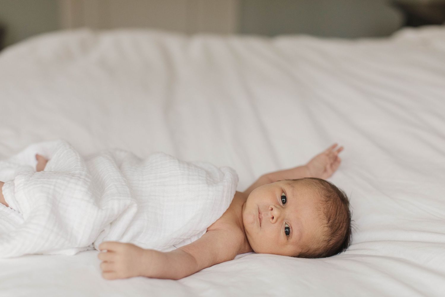 Infant wearing white onesie lying peacefully on white bedding.