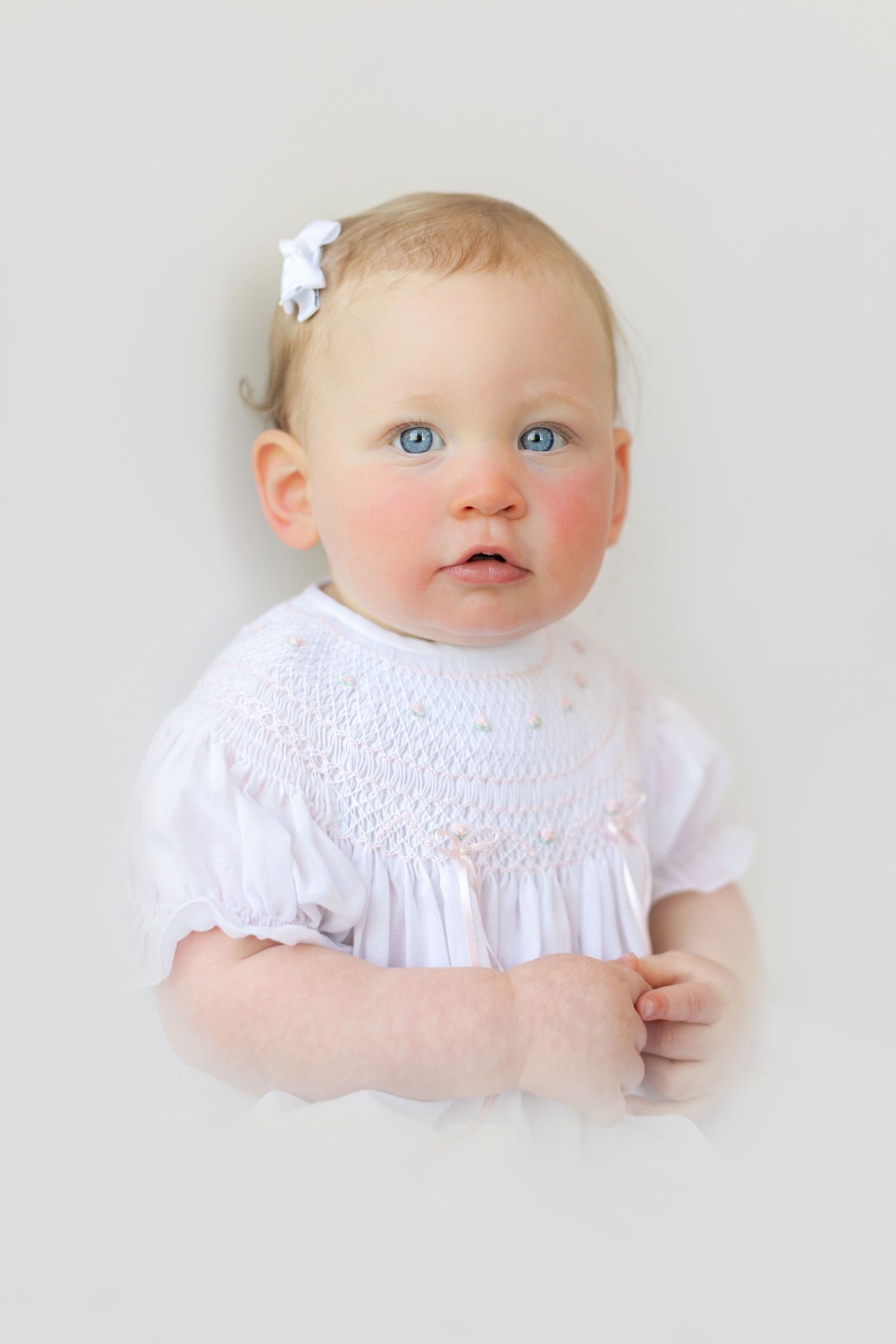 A blue-eyed baby girl in a white smocked dress with a white bow in her blonde hair poses for a portrait.