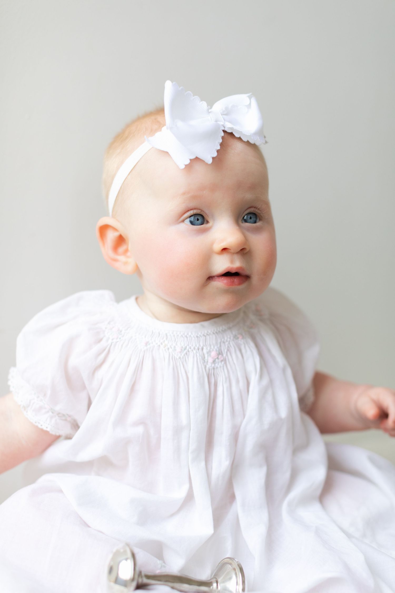 Adorable baby girl with blue eyes wearing a white smocked dress and white bow headband, sitting against a light background.