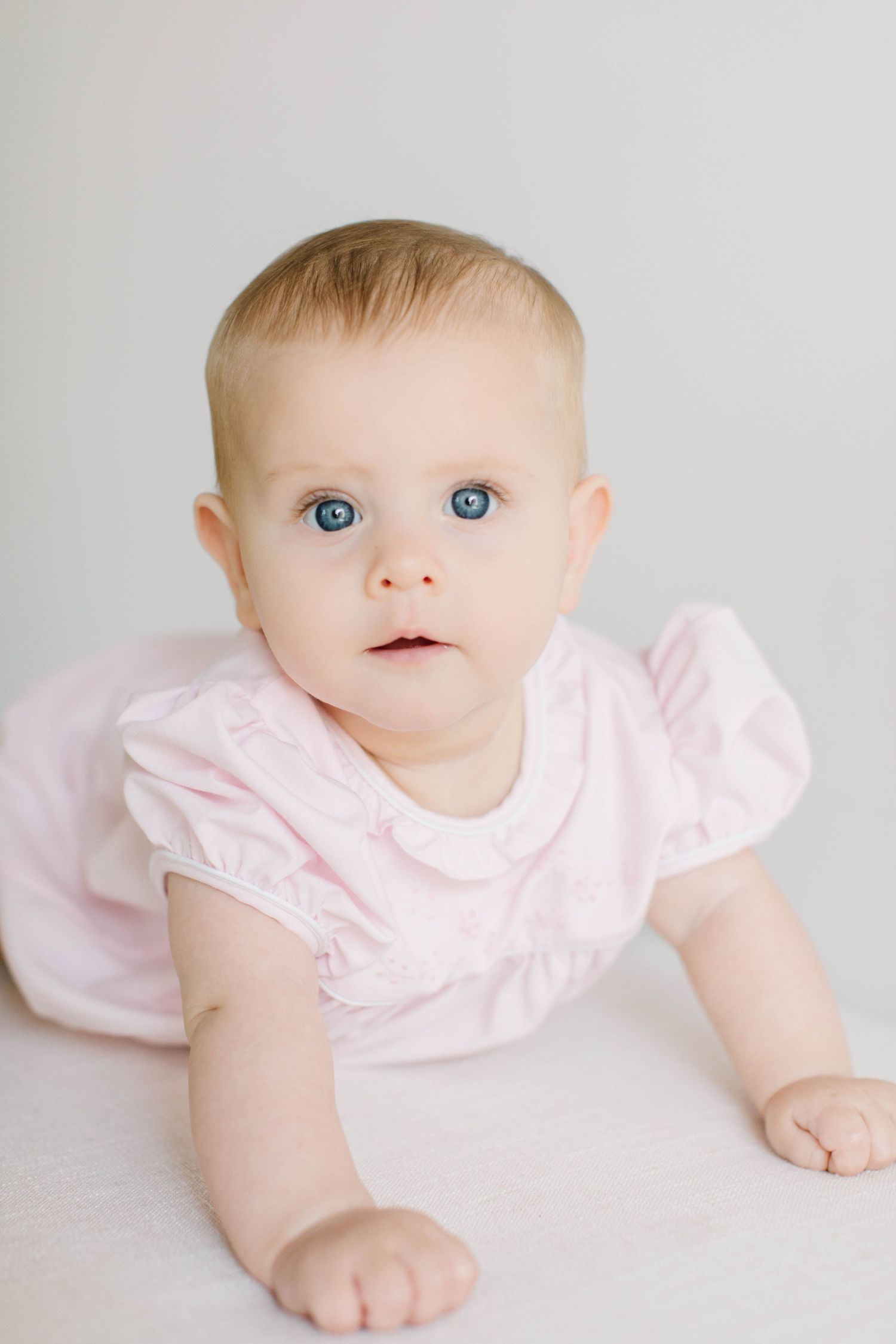 Baby in pink ruffled dress crawls while looking up with bright blue eyes against light background.