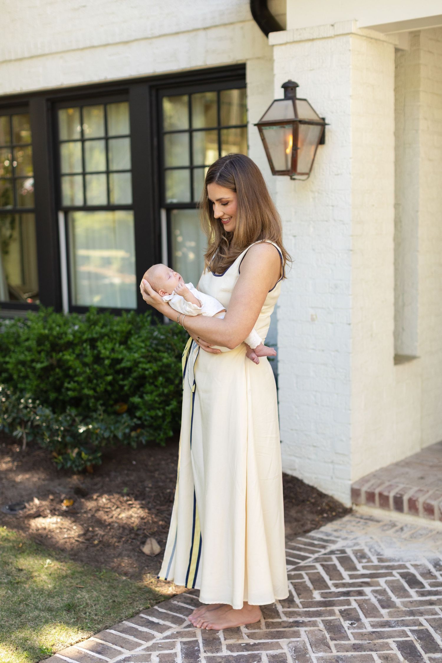 New mother in cream dress tenderly holding newborn baby outside elegant white brick home.