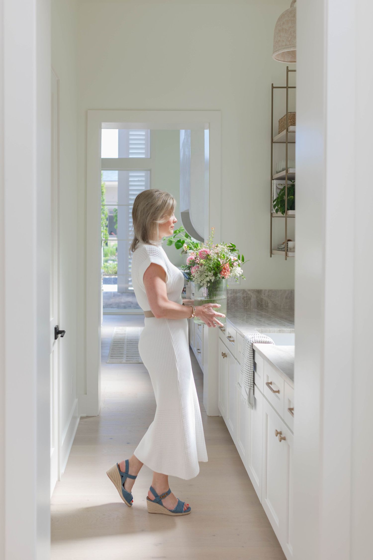 Woman in white dress arranging fresh flowers on a marble kitchen counter in a bright, elegant white kitchen.