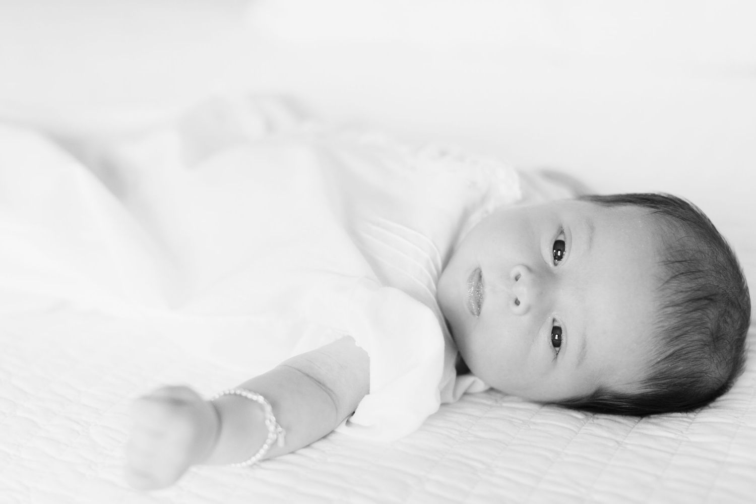 Black and white photo of a peaceful newborn baby lying on a white blanket, gazing softly at the camera.