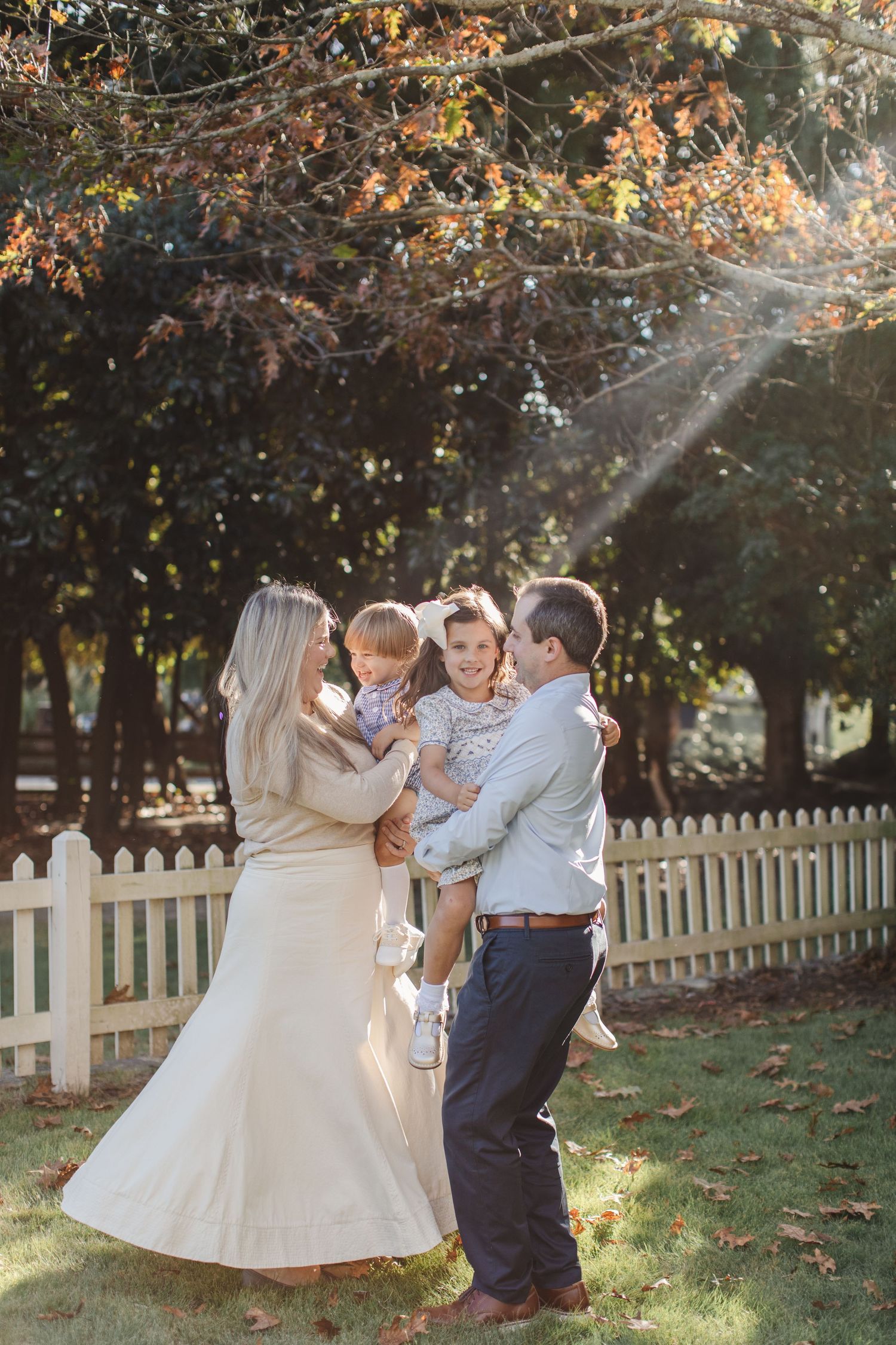 Children in formal attire dance and play together in a backyard with autumn trees and a white picket fence in the background.