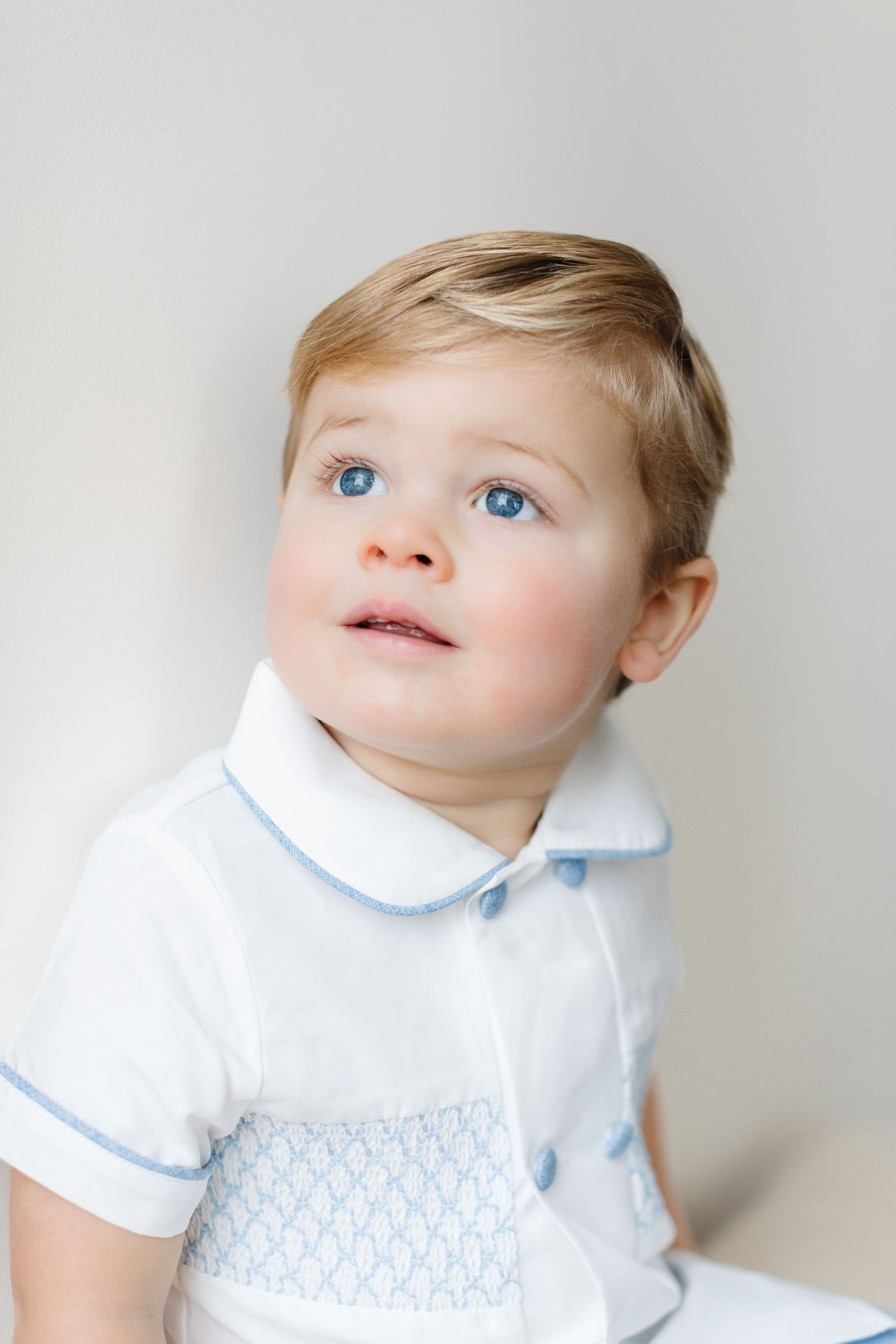 A toddler in a white collared outfit with blue trim looks up with bright blue eyes.
