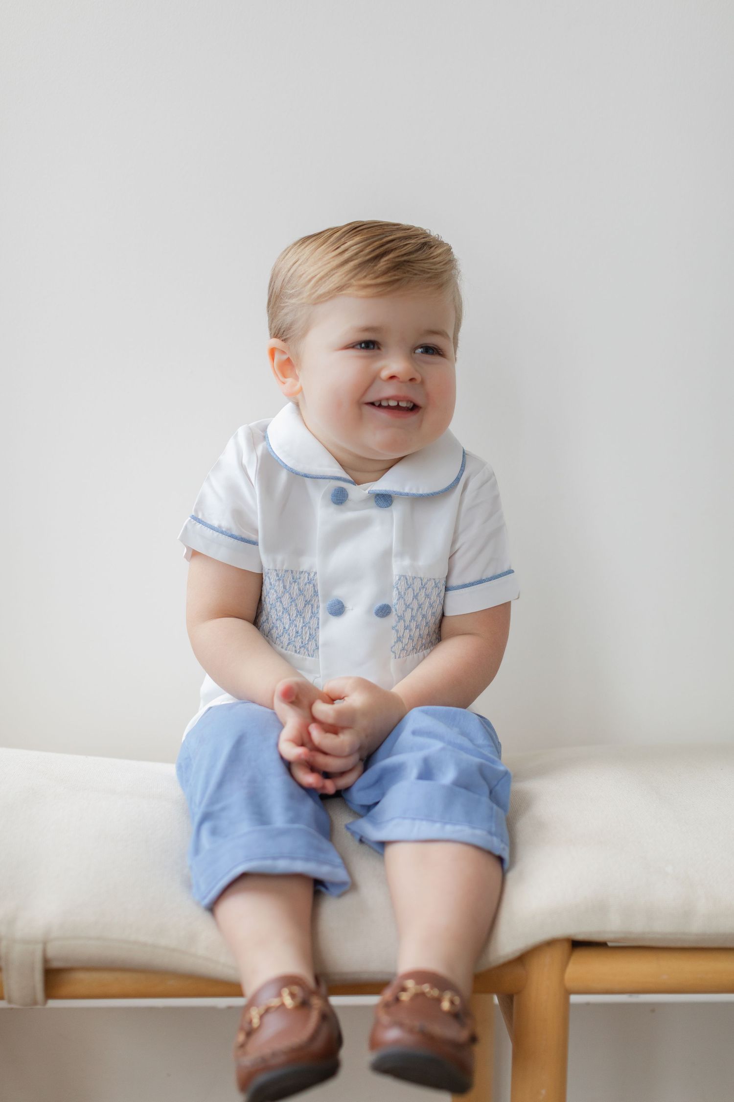 Sweet baby wearing light blue shorts and white collared shirt sits on wooden bench against white wall.