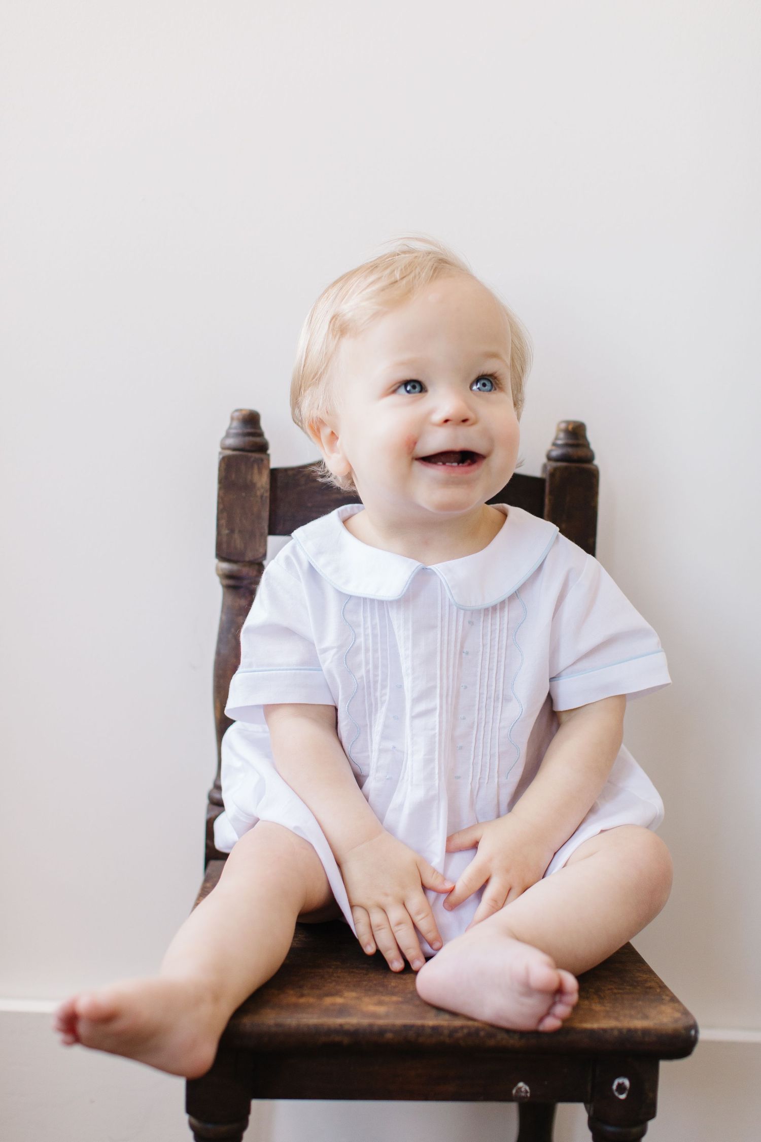 Smiling baby in white dress sitting on vintage wooden chair against white wall.