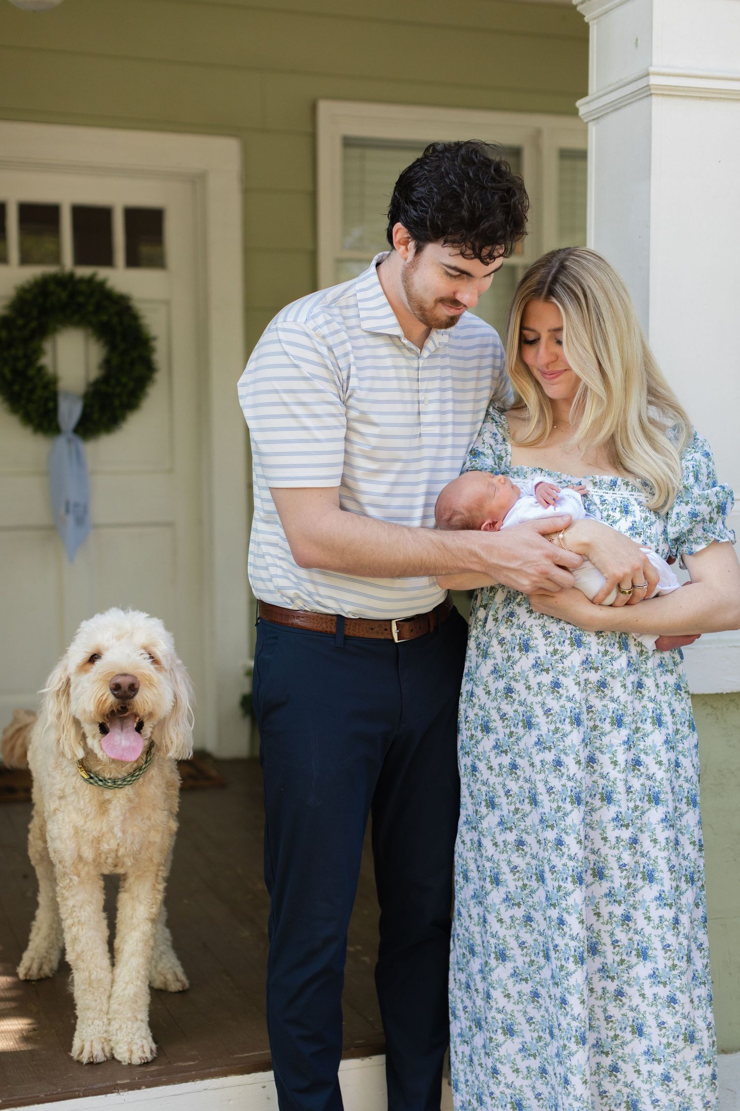 Young couple with curly-haired man and blonde woman in floral dress holding newborn baby beside fluffy Goldendoodle on porch.