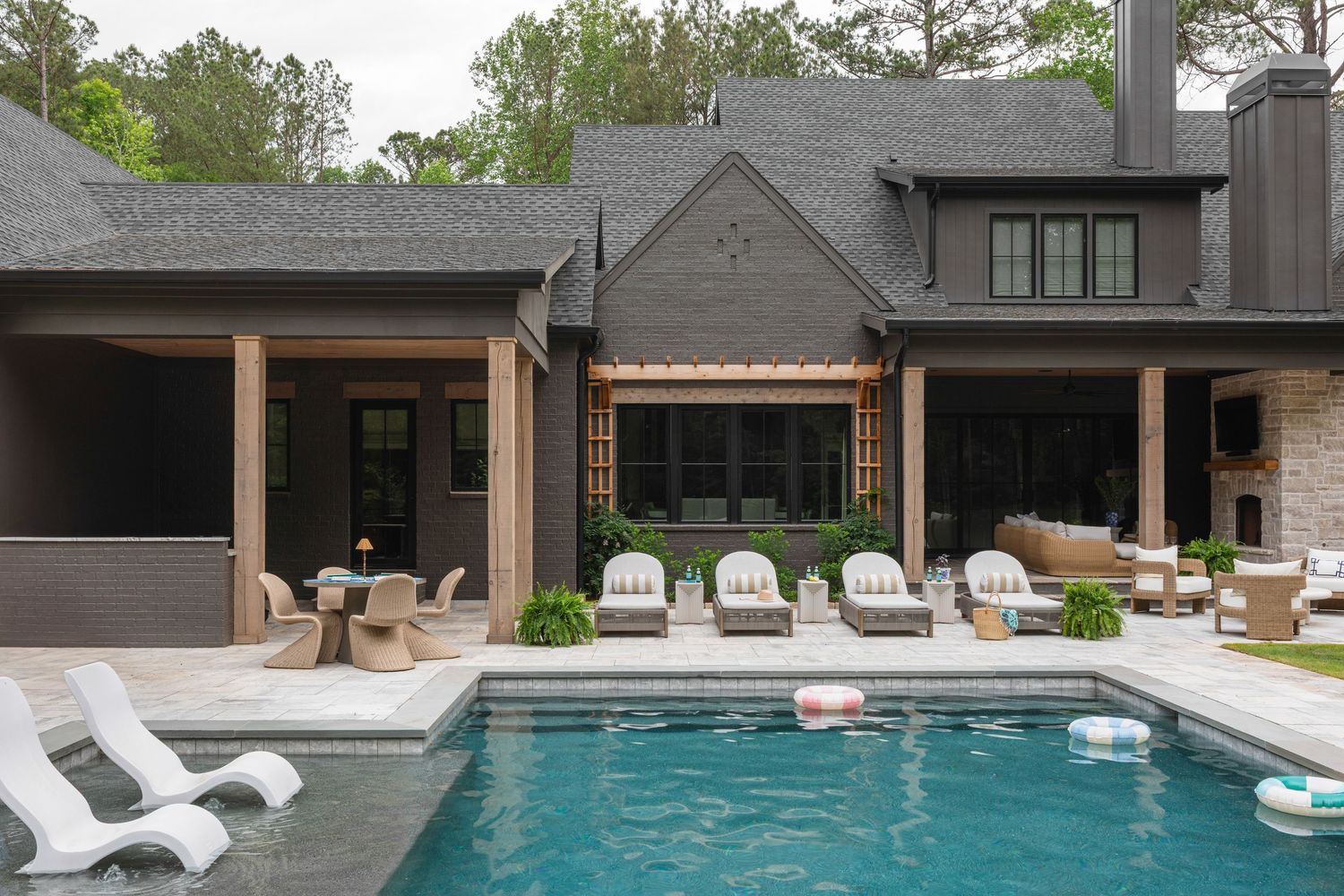 Elegant backyard pool area with lounge chairs, covered patio, and lush landscaping surrounding a dark-painted luxury home.