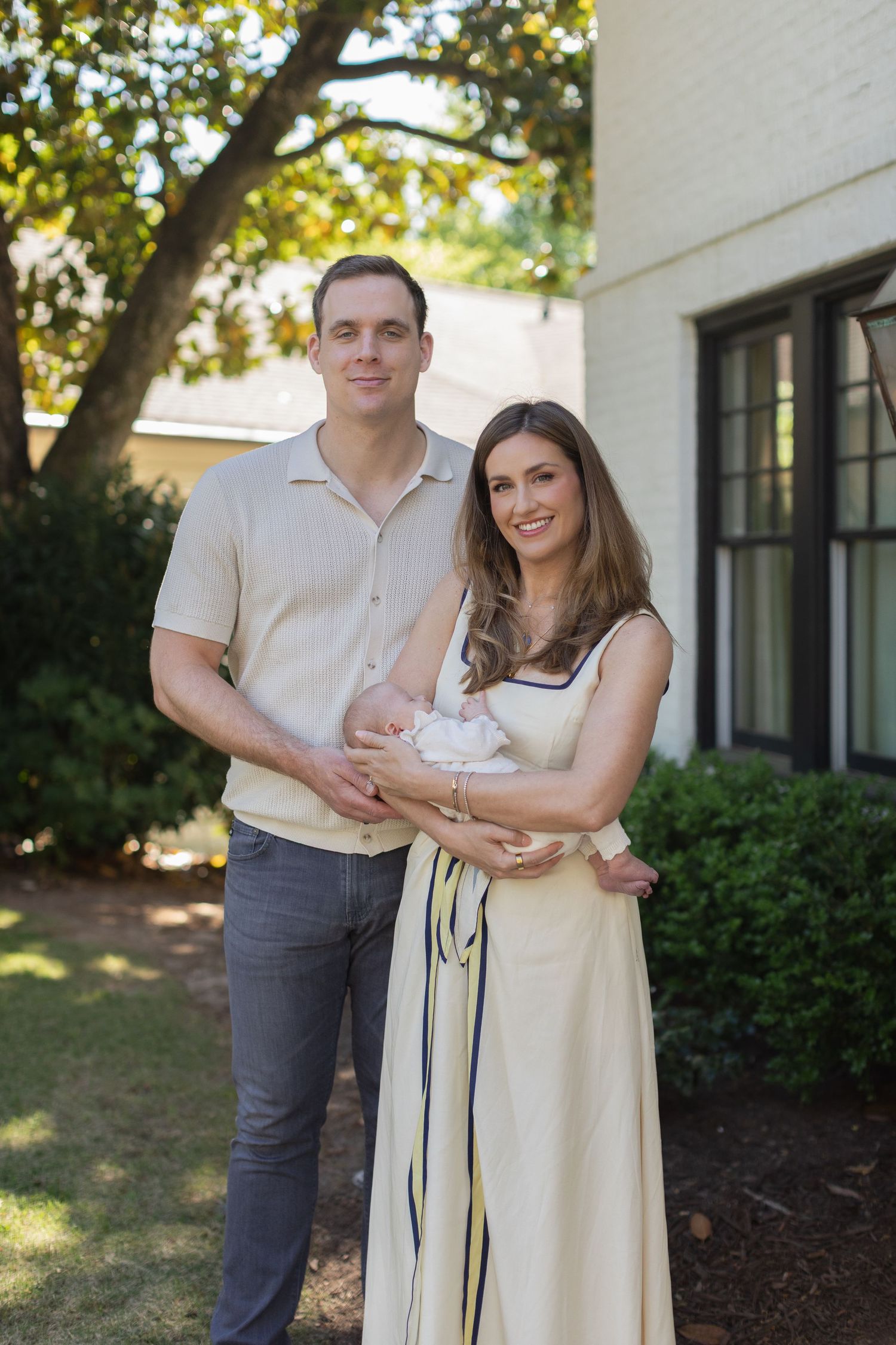 Happy couple posing outdoors holding their newborn baby in front of a white brick home.