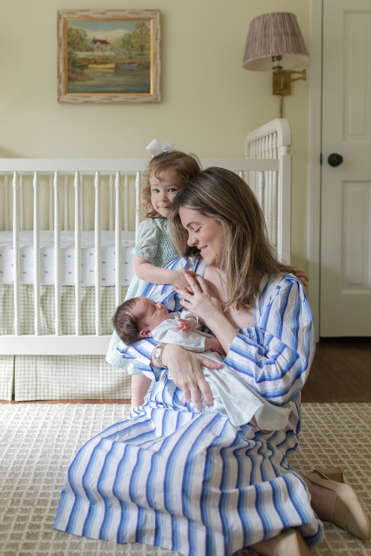 A tender moment in a nursery with a striped blue dress, white crib, and vintage landscape painting on the wall.