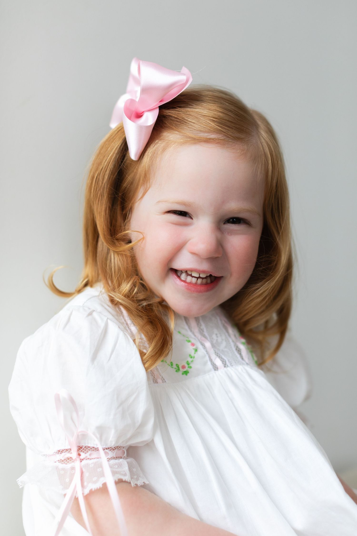 Smiling redhead toddler girl wearing white dress and pink bow hair clip, cheerful portrait photo.