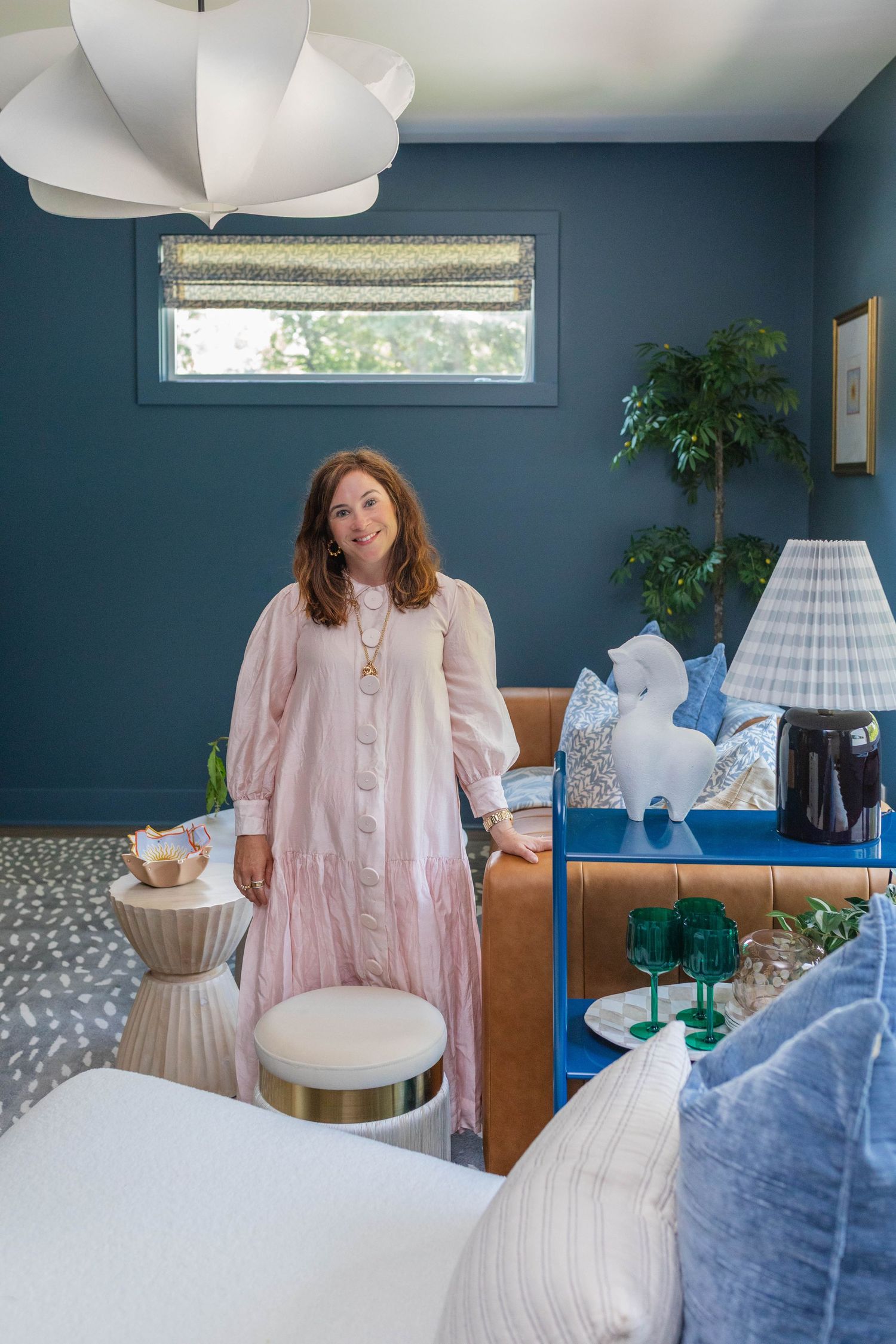 Woman in pink dress stands in a stylish blue bedroom with camel sofa, white pendant light, and chic decor accents.