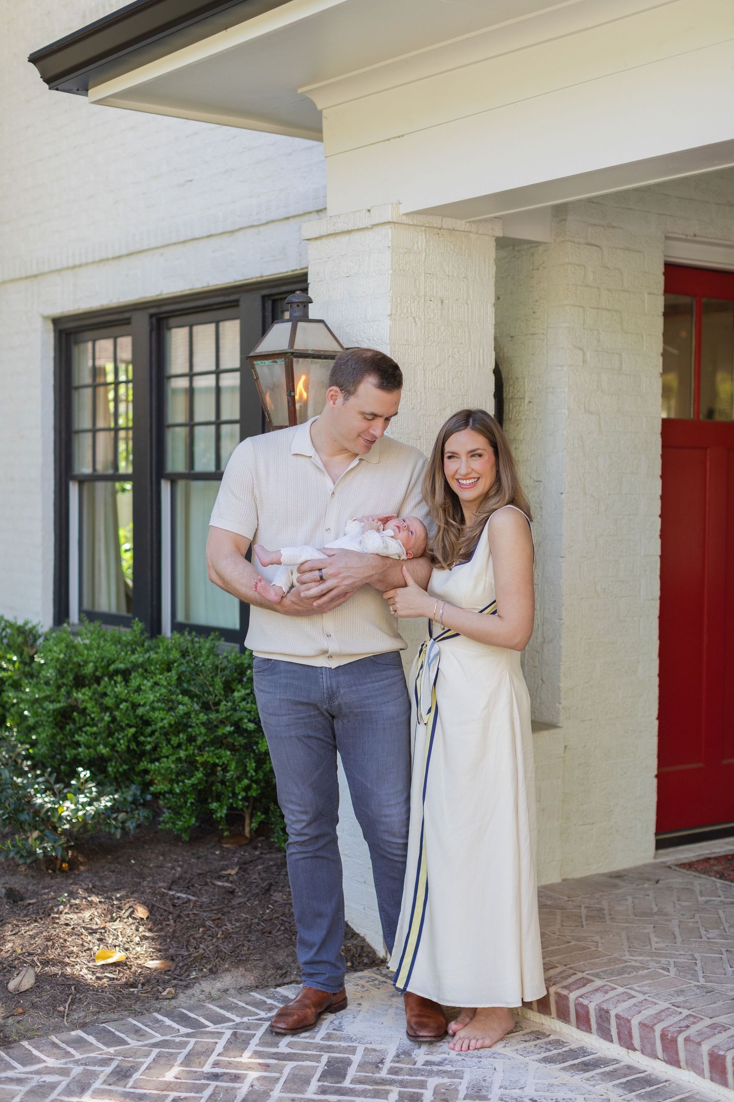 New parents stand outside their white brick home holding their newborn baby, dressed in neutral tones.