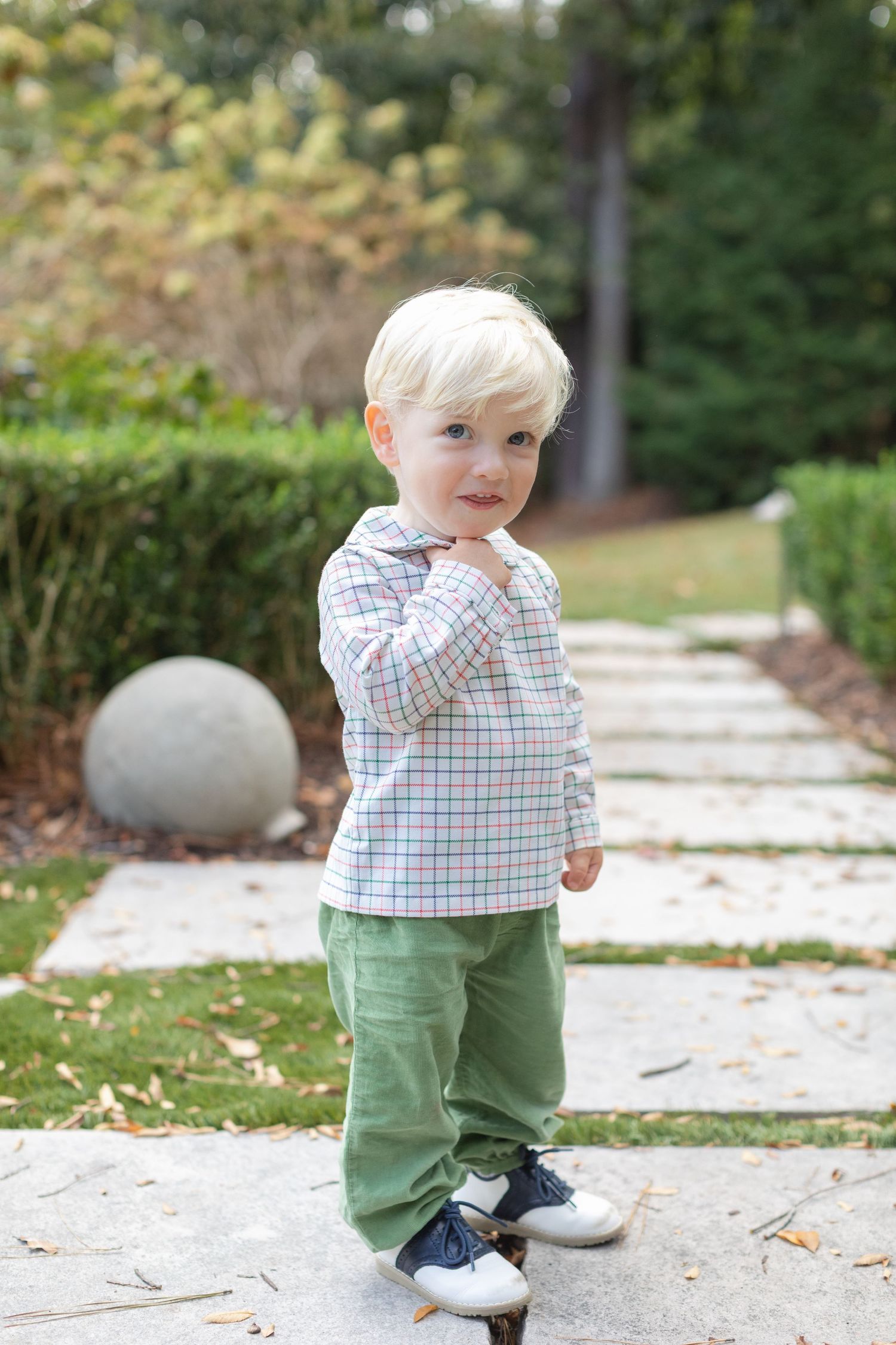 A toddler in green pants and plaid shirt walks along a stone path on an autumn day.