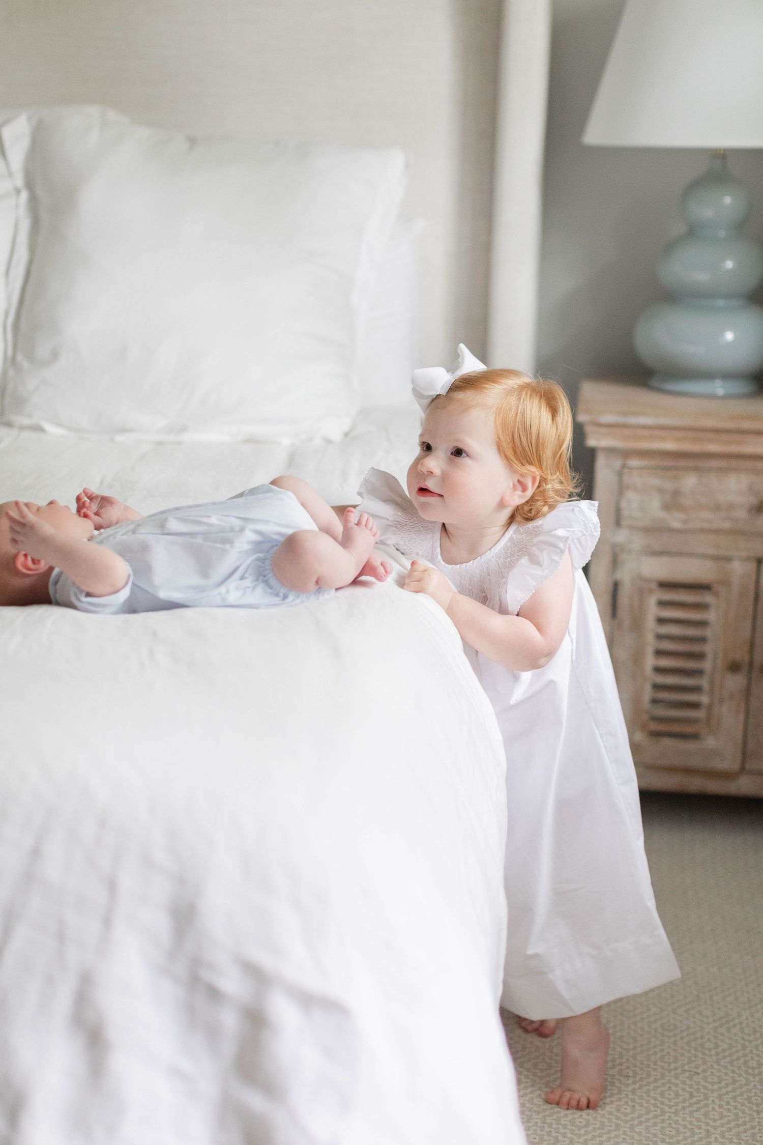 A toddler in a white dress stands beside a bed with white linens and looks upward with joy.