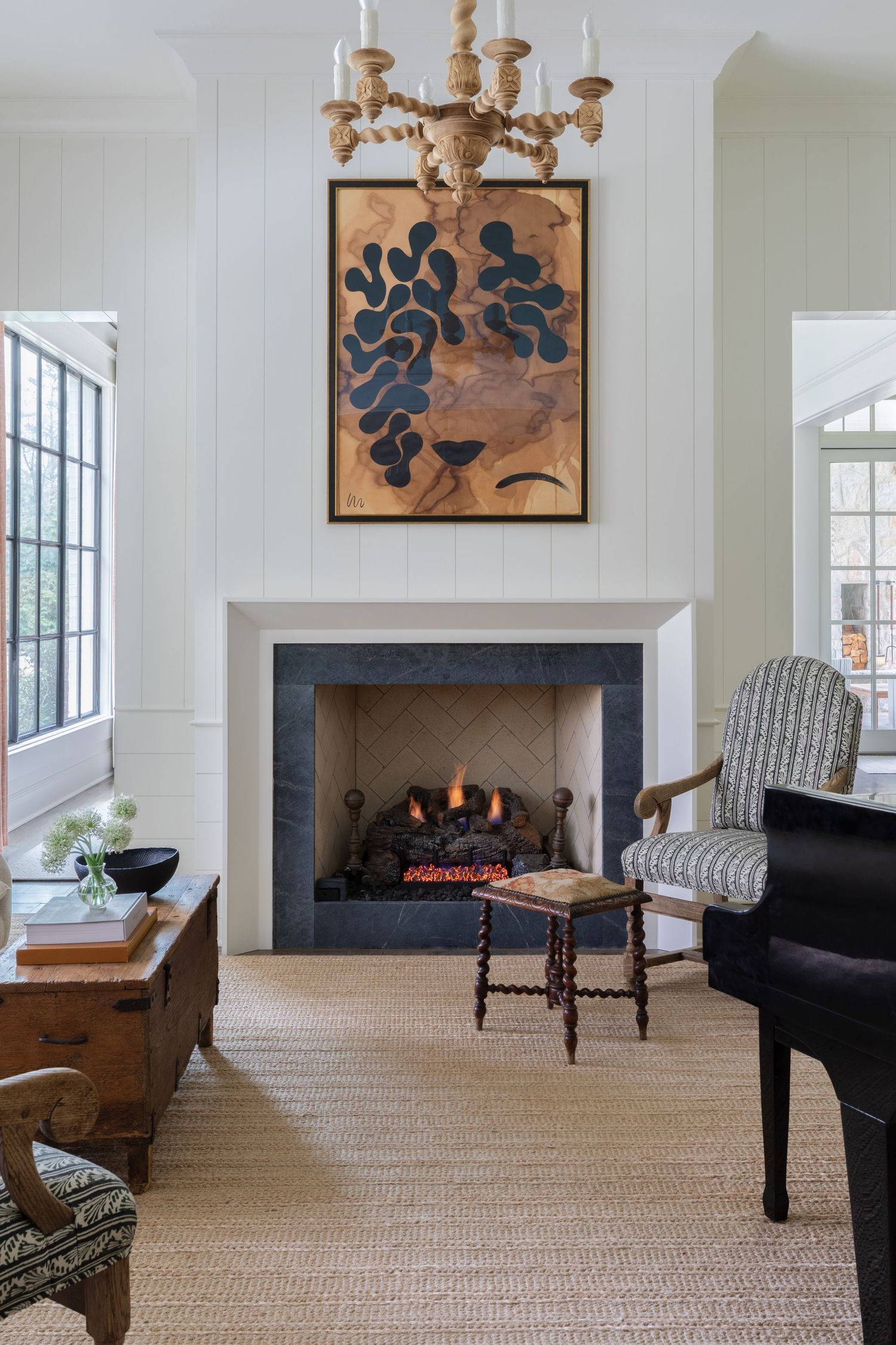 Cozy living room with lit fireplace, botanical artwork, chandelier, patterned armchair, and natural fiber rug.