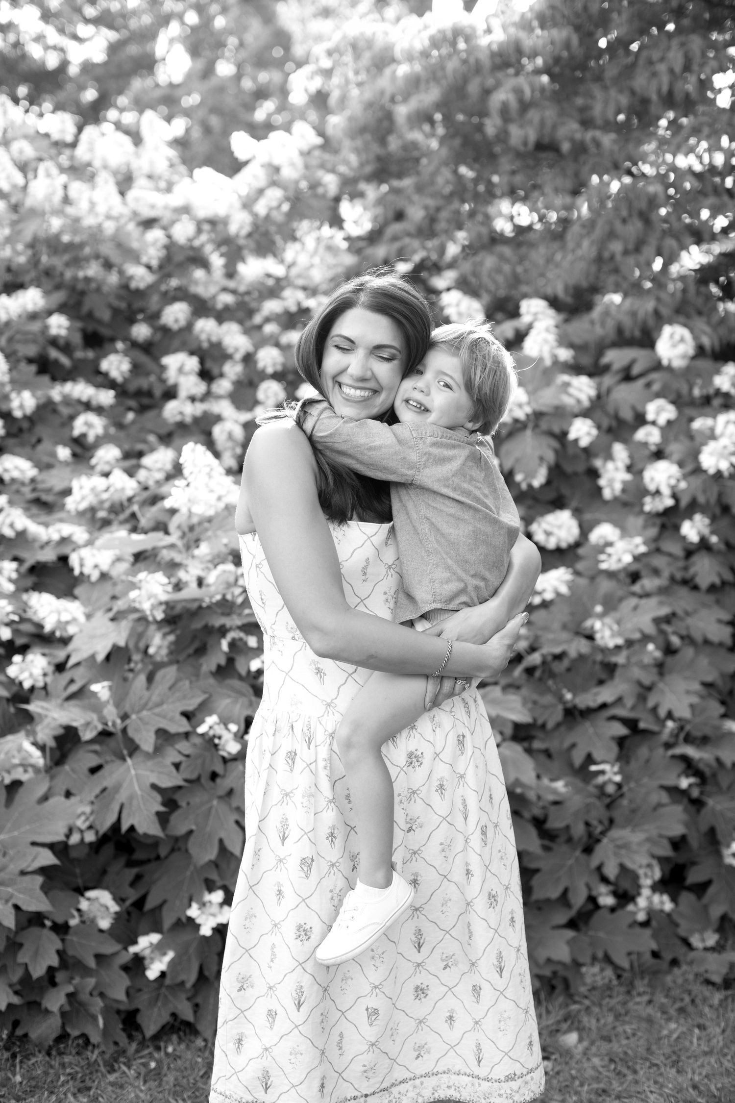 Black and white photo of a smiling mother holding her laughing child in front of blooming hydrangea bushes.
