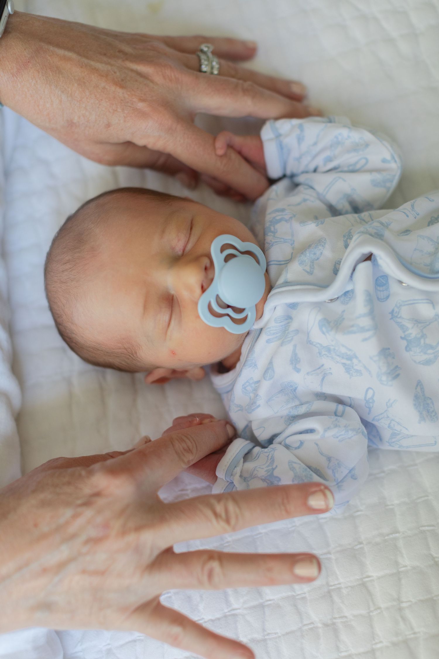 Sleeping newborn baby in blue outfit with pacifier resting peacefully in crib with adult hands nearby.
