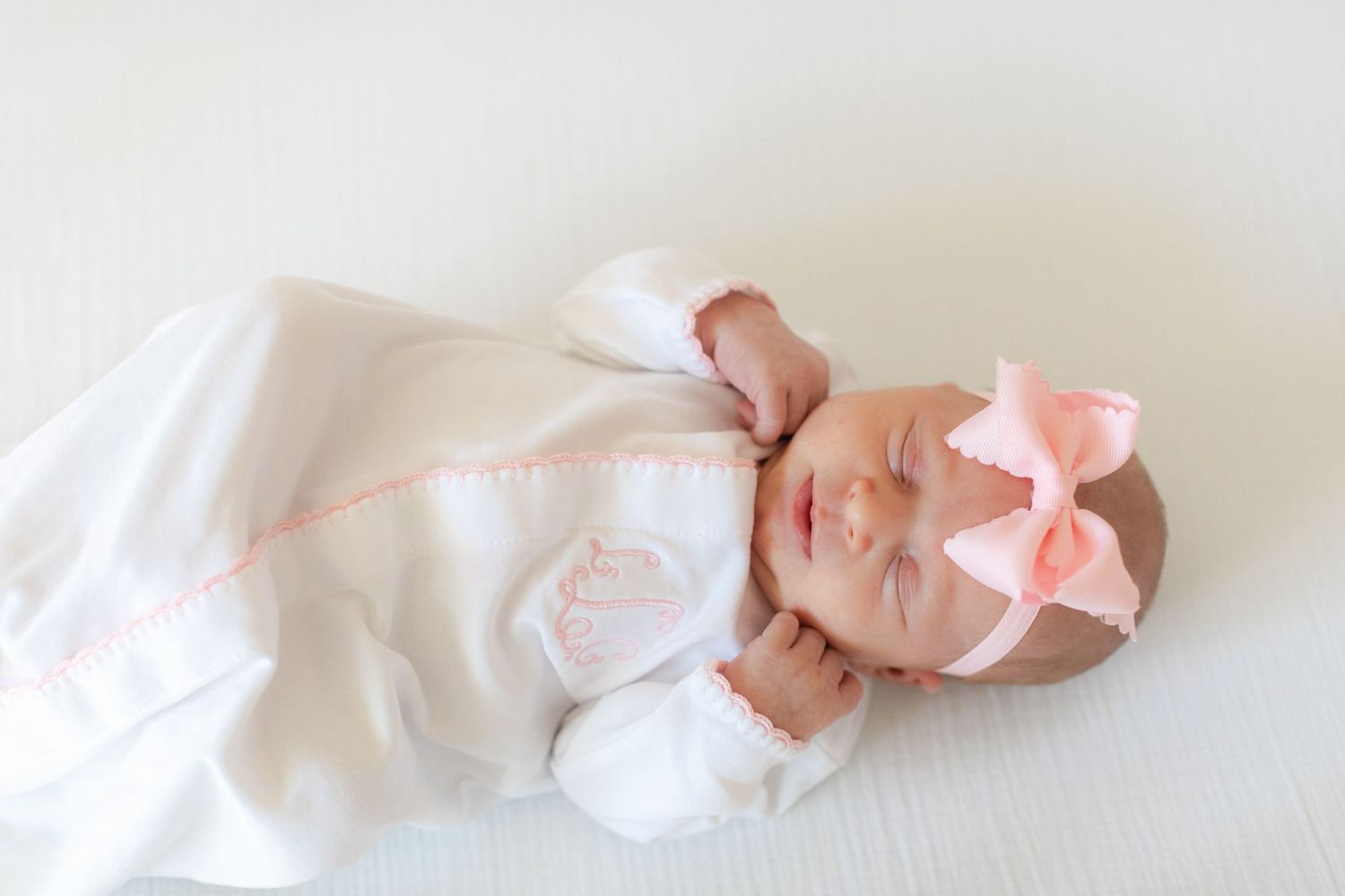 A newborn baby wearing a white outfit and pink bow sleeps peacefully in a white blanket.