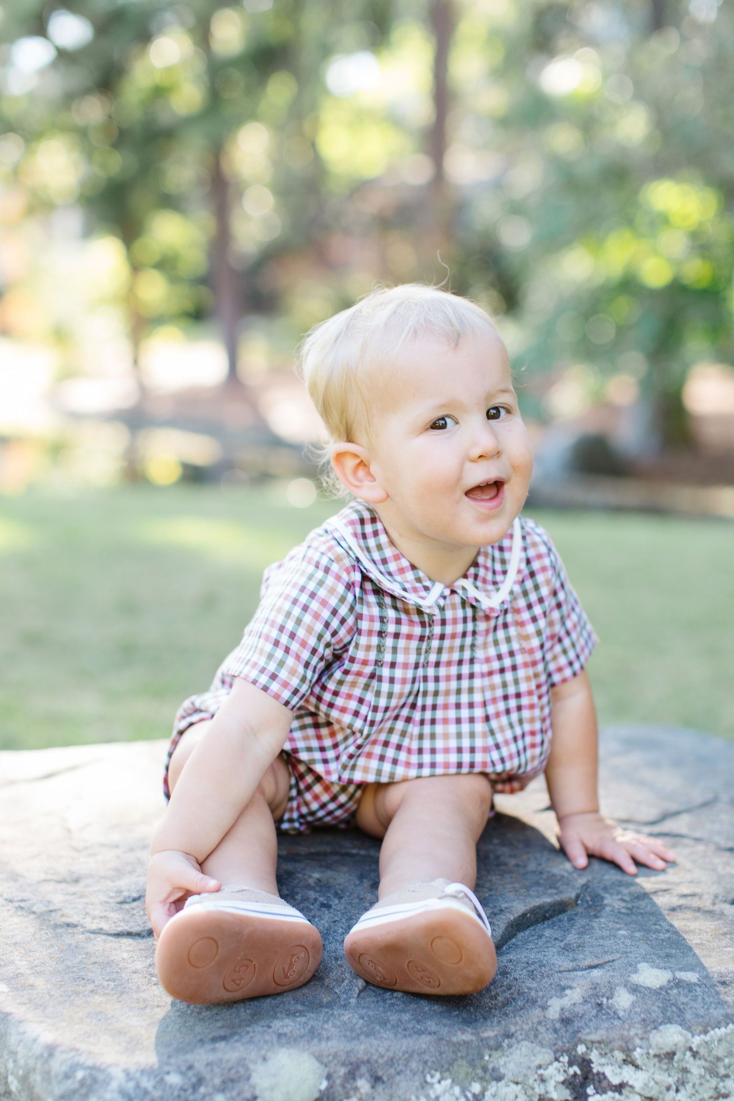 A baby in gingham shirt and denim shorts sits and crawls on a large rock outdoors in a park setting.