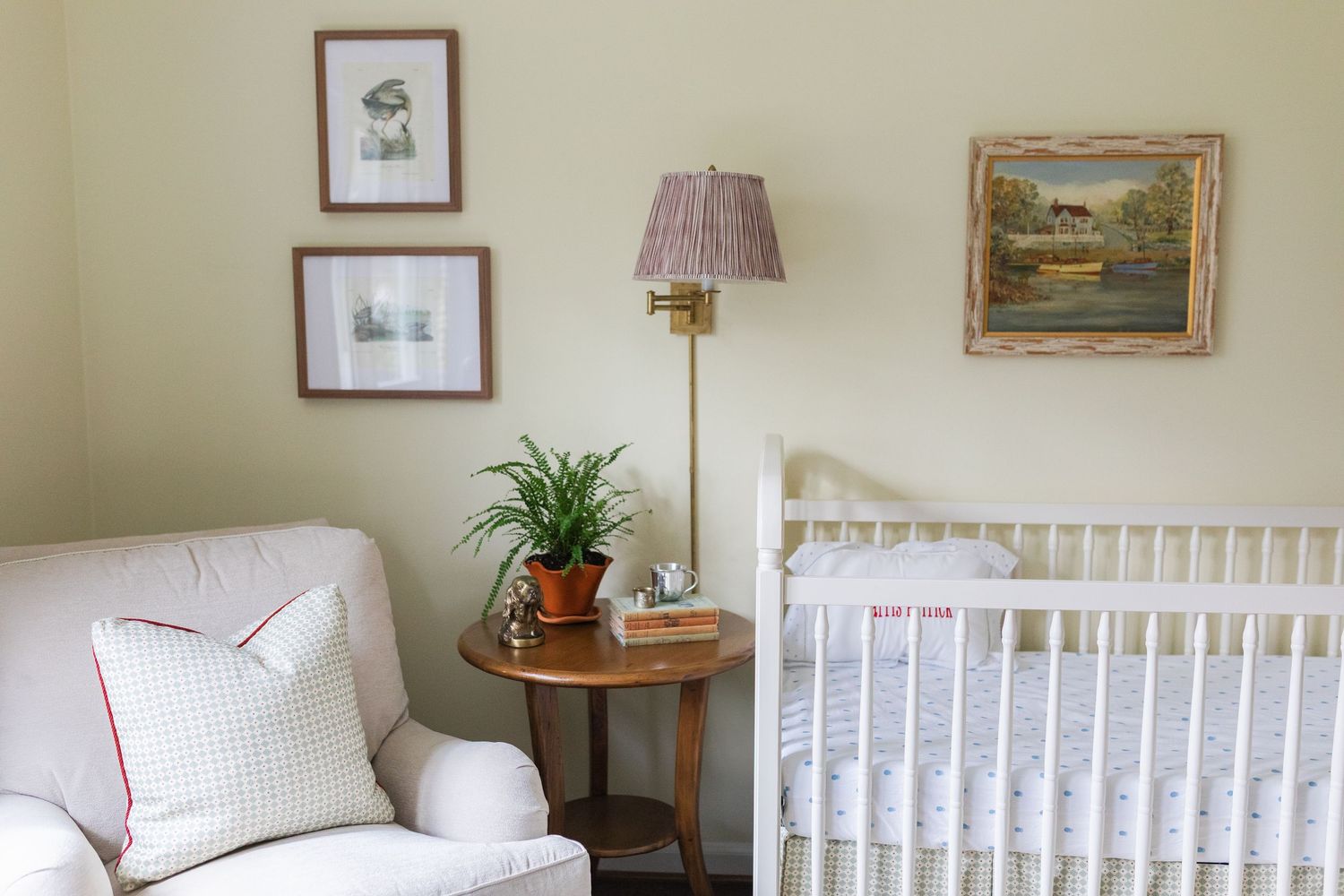 A cozy nursery room with white crib, plush chair, framed artwork, and potted plants creating a serene atmosphere.