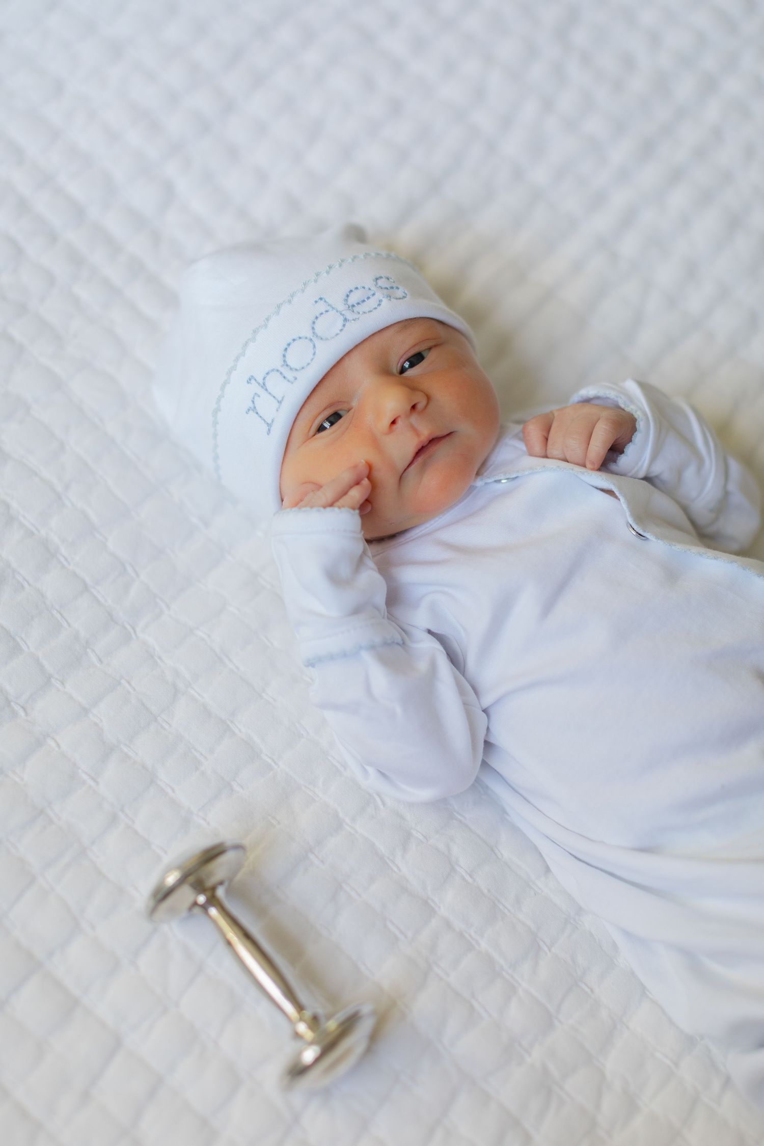 Newborn baby named Rhodes in white outfit and personalized hat lies next to a silver rattle.