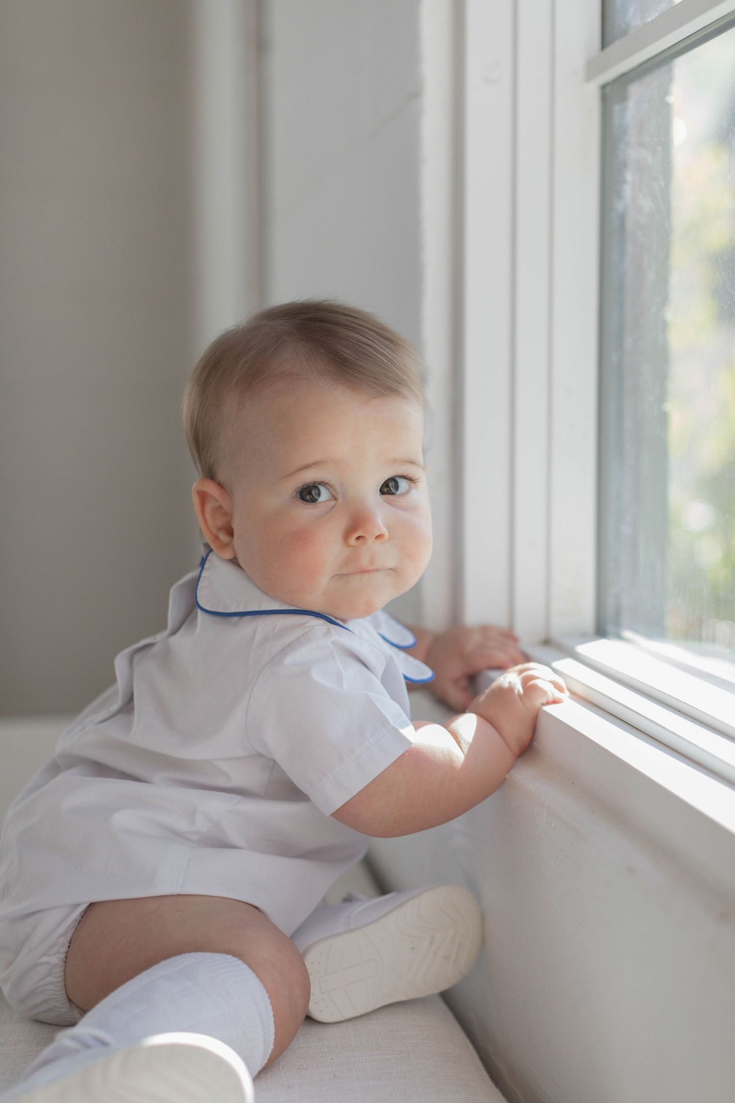 Baby in white romper looking back while sitting at window sill in natural light. Atlanta Children's Fine Art Heirloom Portraits
