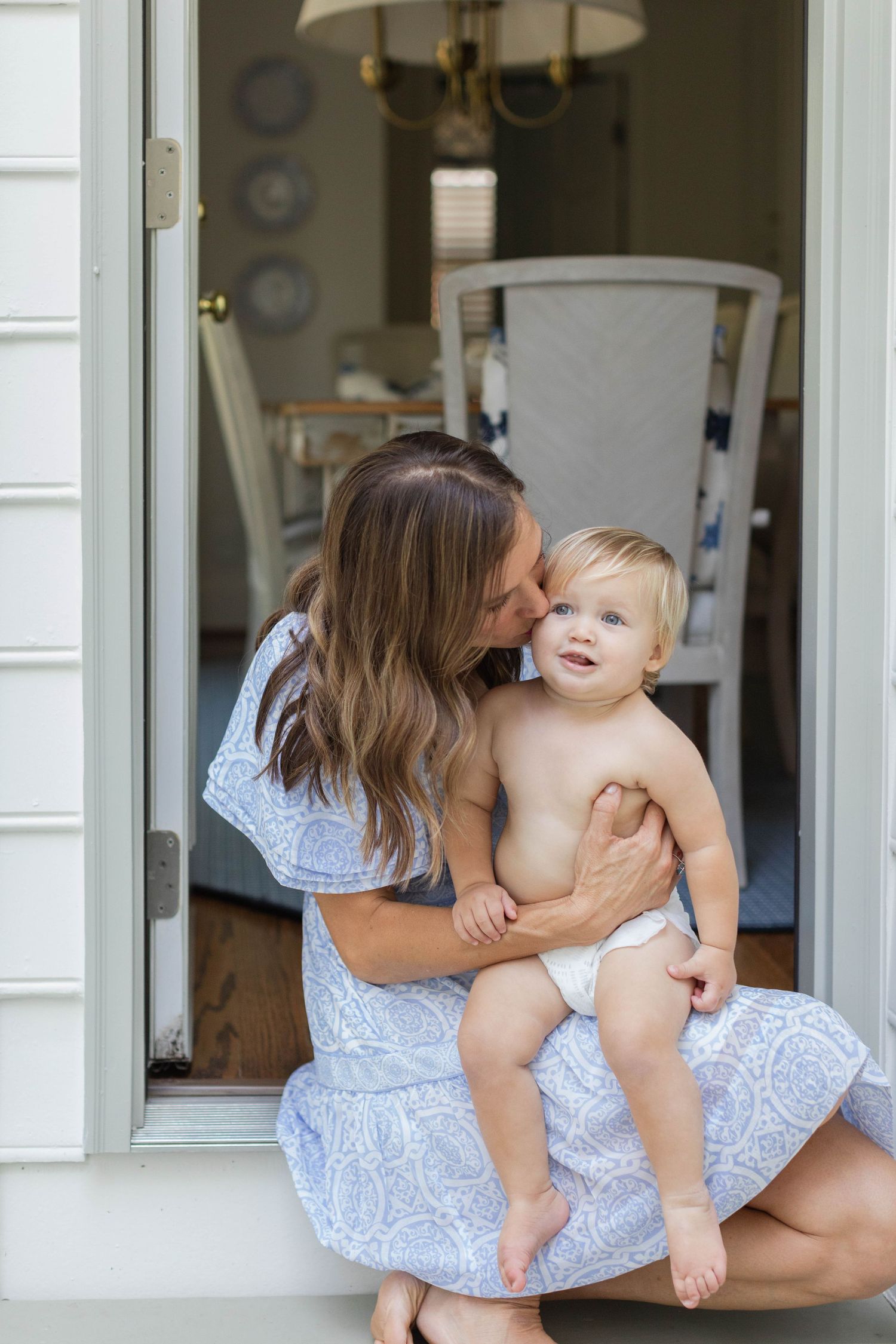 Mother in blue dress playfully holding and kissing baby on front porch in summer.