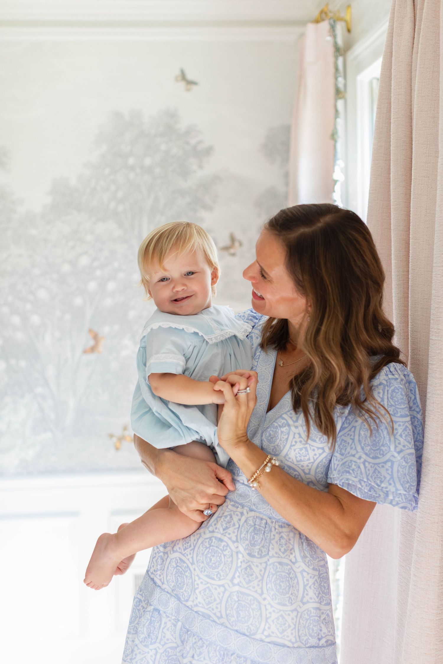 A parent wearing a blue floral dress holds and cuddles with a baby in white against a light background.