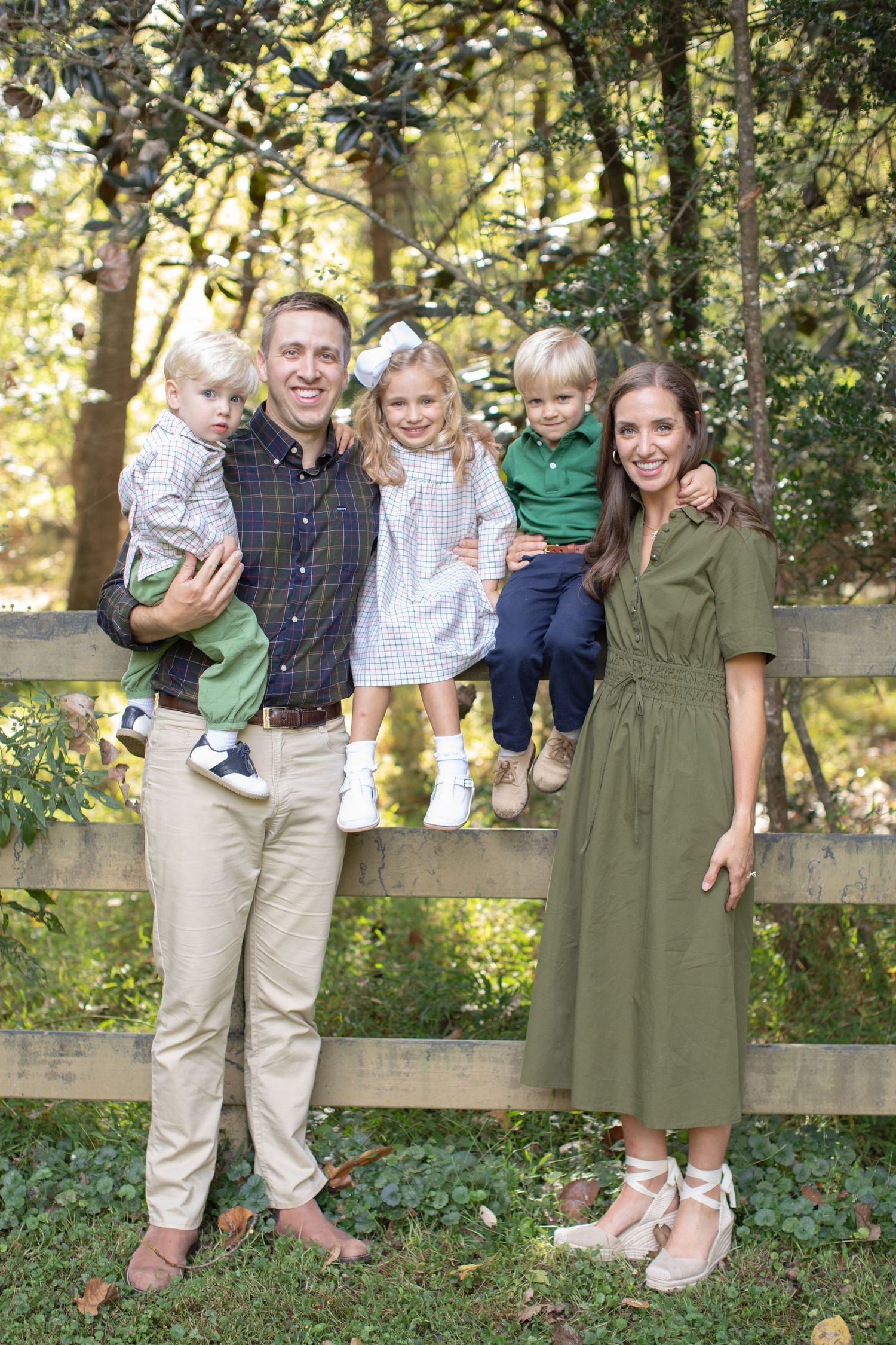 A family poses together by a wooden fence in a wooded setting during autumn.