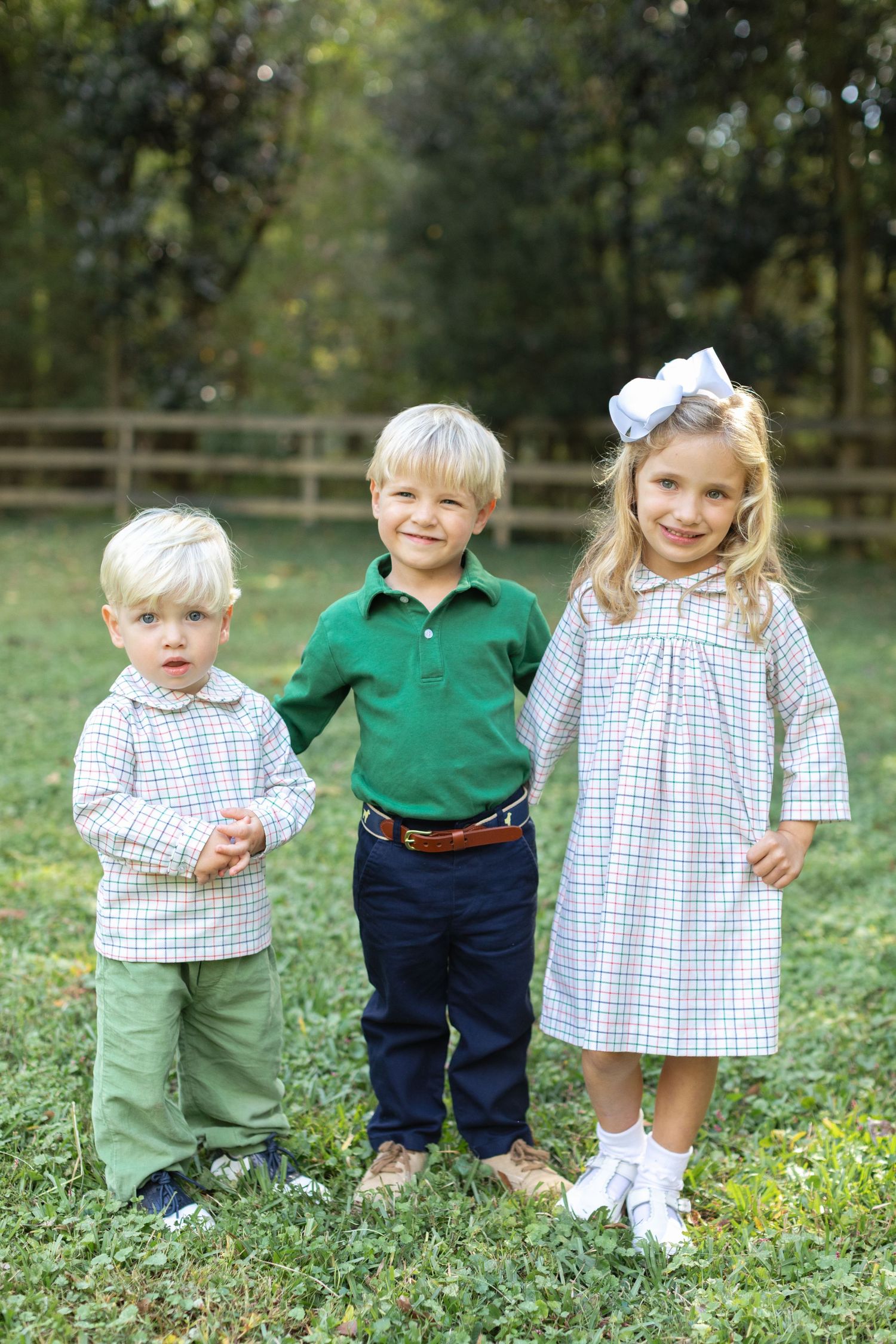 Three young children in coordinated outfits play together in a grassy field with flowers on a sunny day.