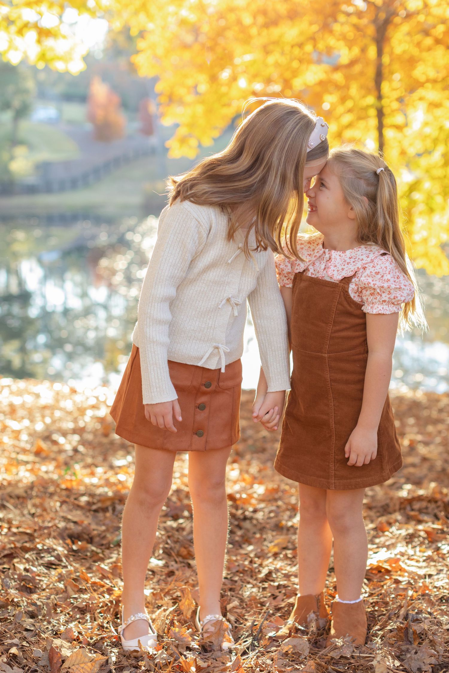 Two children in brown corduroy dresses share a sweet moment among golden autumn leaves on a sunny fall day.