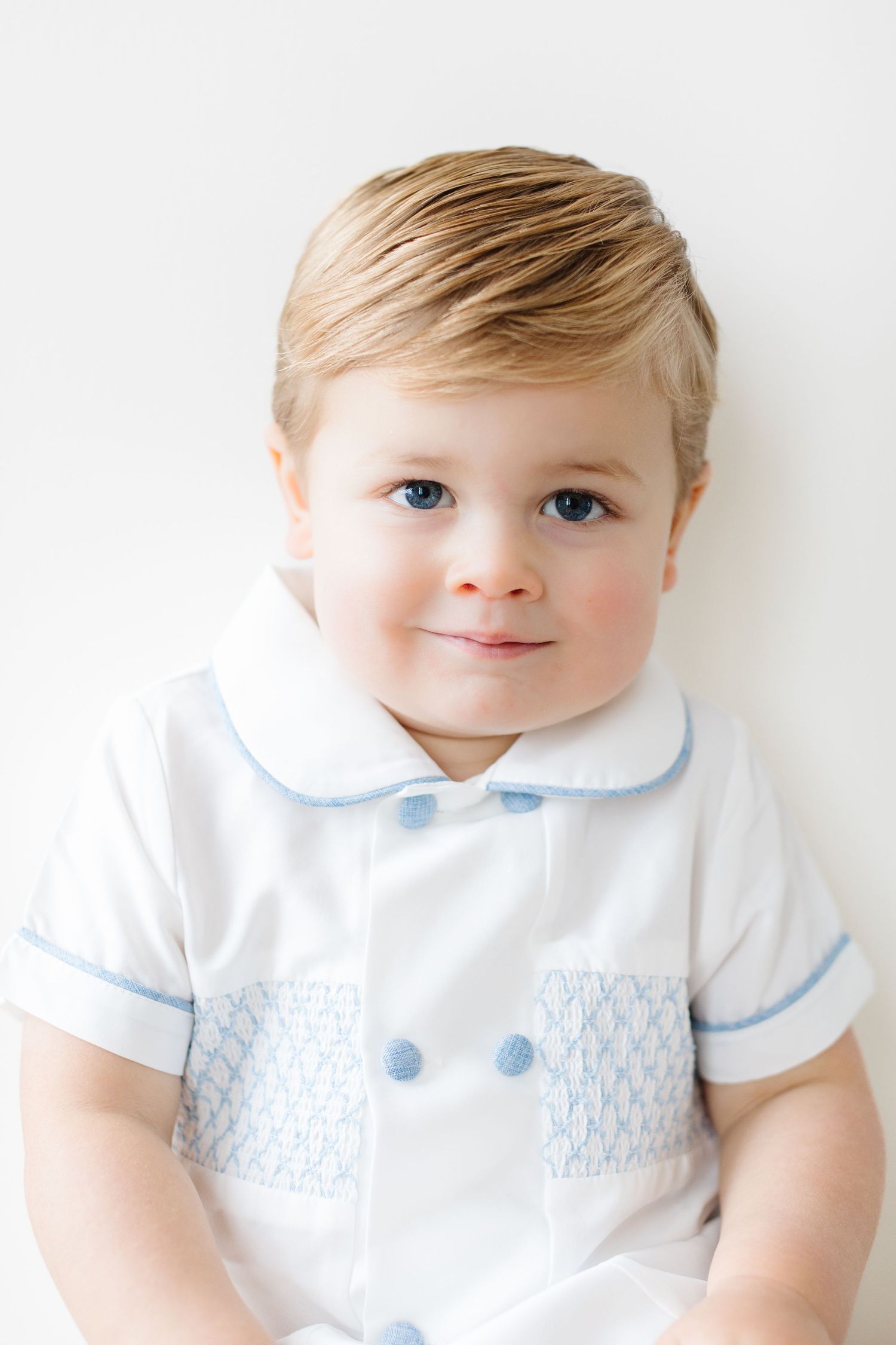 Adorable toddler wearing white peter pan collar outfit with blue smocking detail smiles at camera.