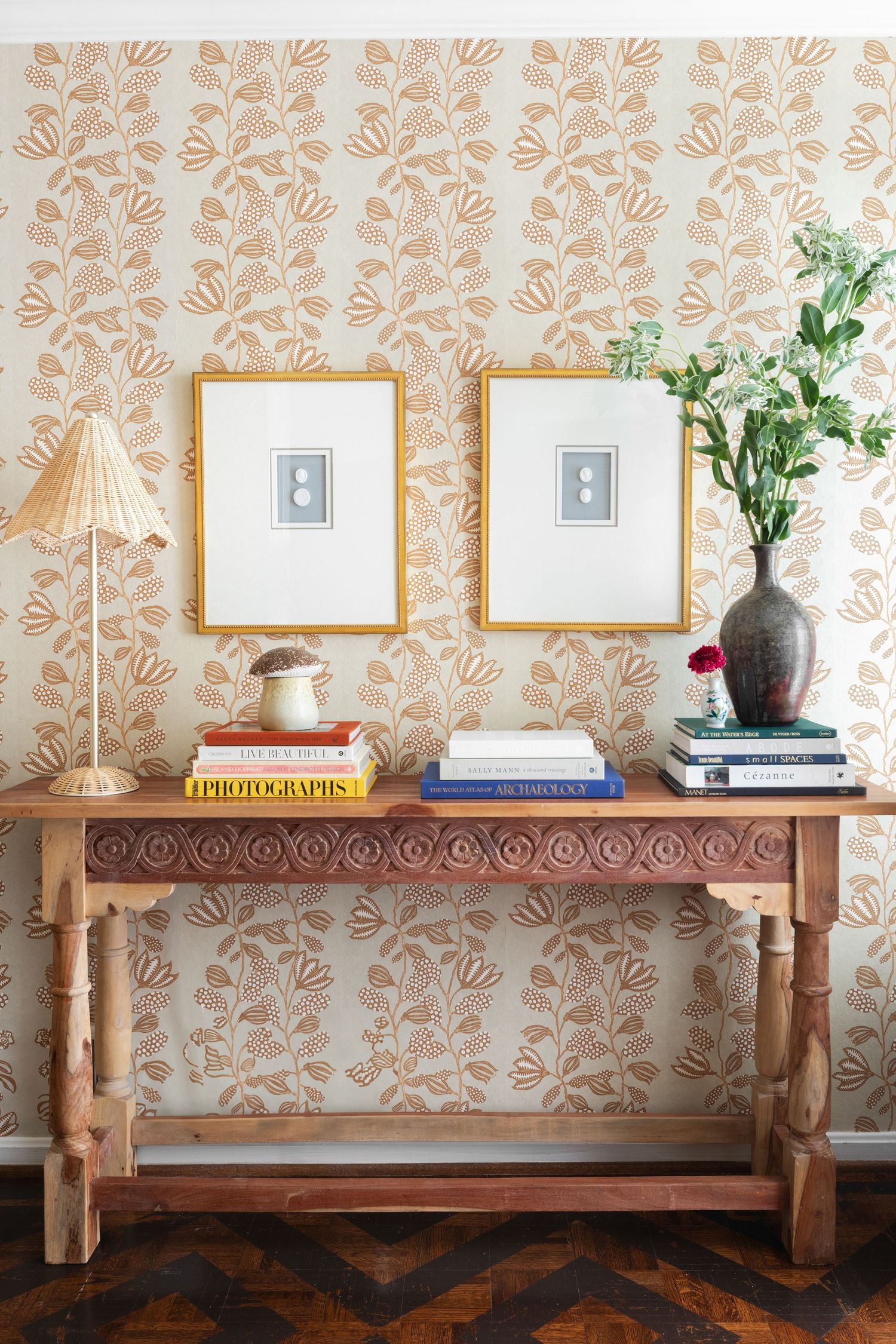 Wooden console table with carved details, stacked books, vase, and two gold-framed prints against floral wallpaper.