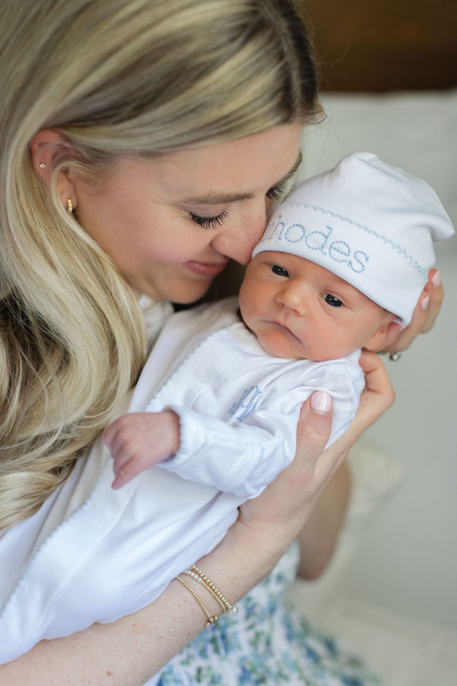 A blonde mother tenderly holds her newborn baby named Rhodes, dressed in a white outfit and personalized hat.