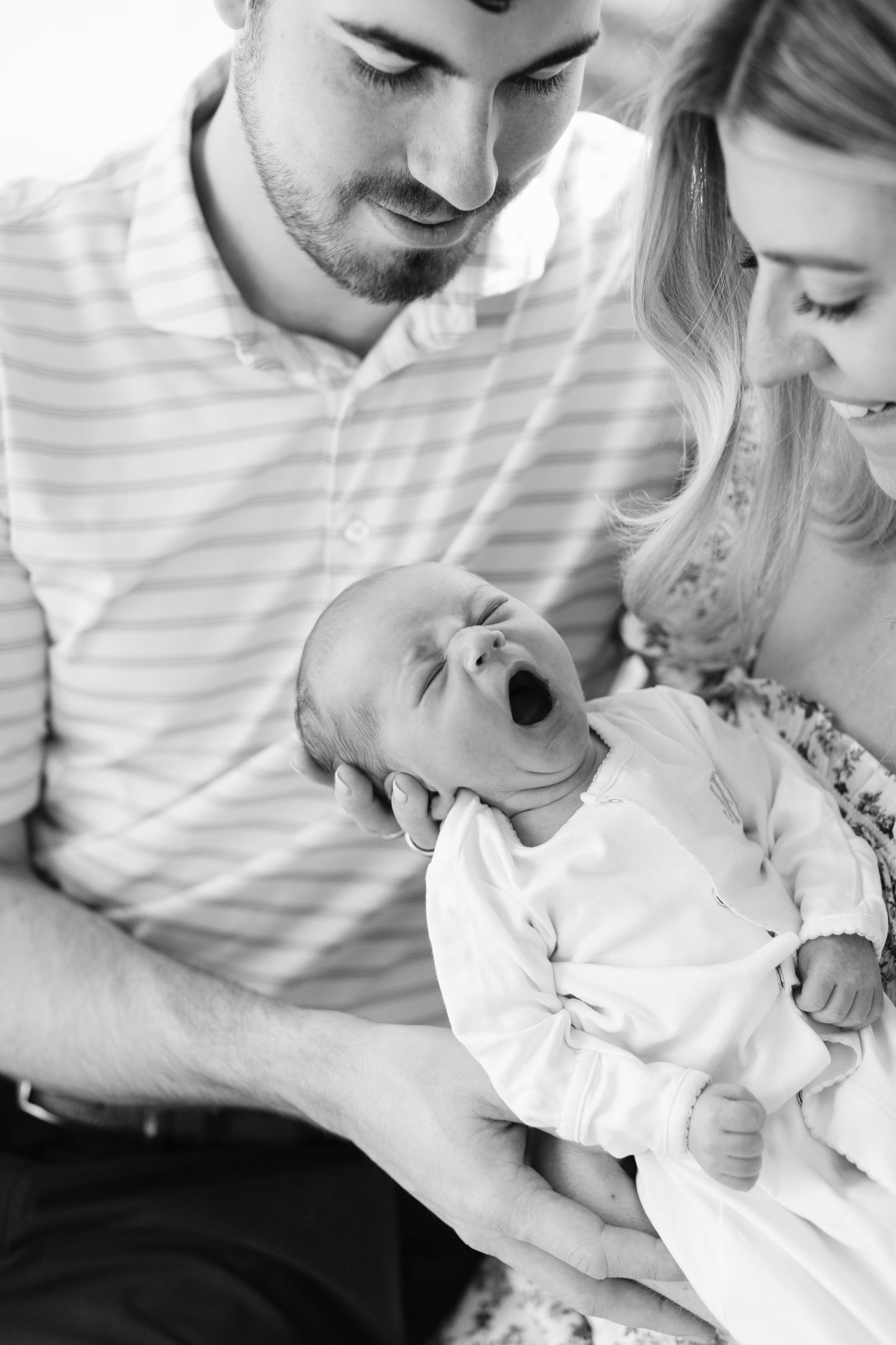 New parents lovingly hold their yawning newborn baby in black and white portrait photo.