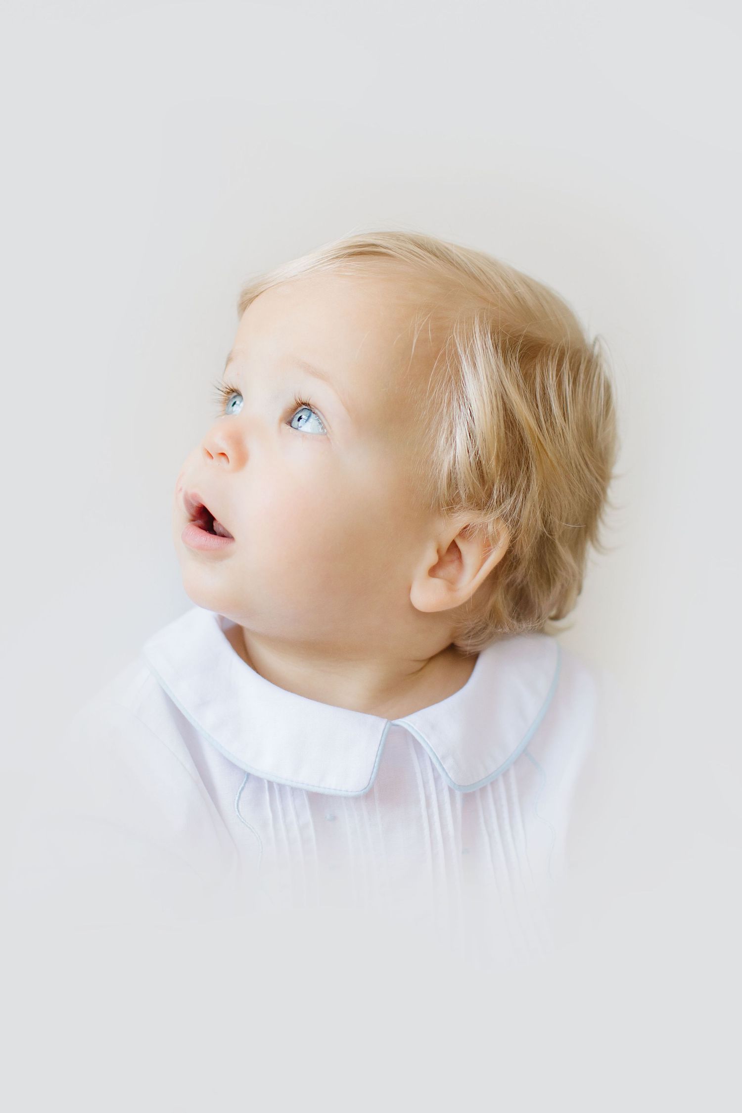Three profile views of a baby with blonde hair wearing a white collar looking upward against a light background.