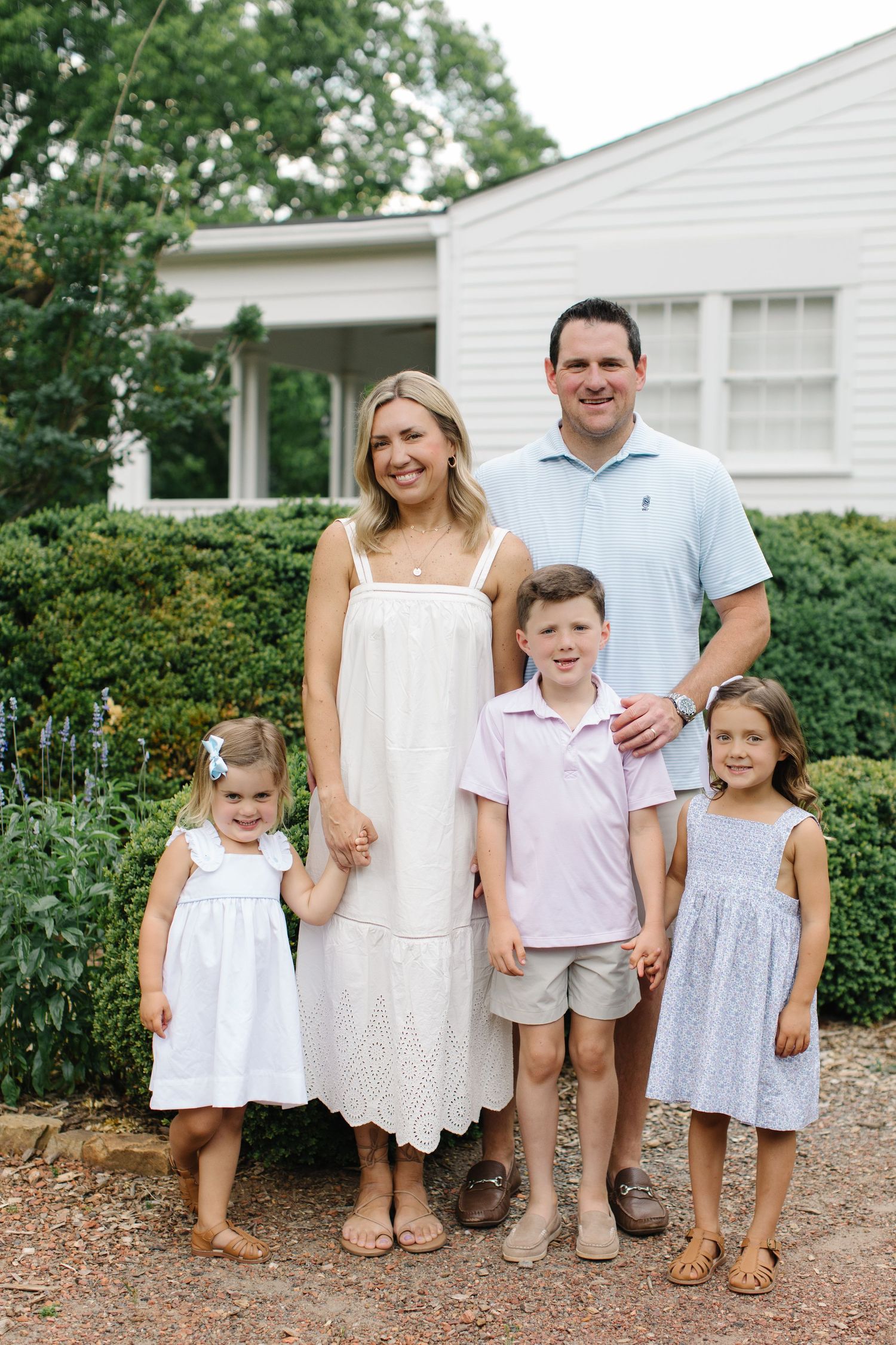 A happy family of five poses outdoors in coordinating pastel outfits in front of a white house surrounded by greenery.