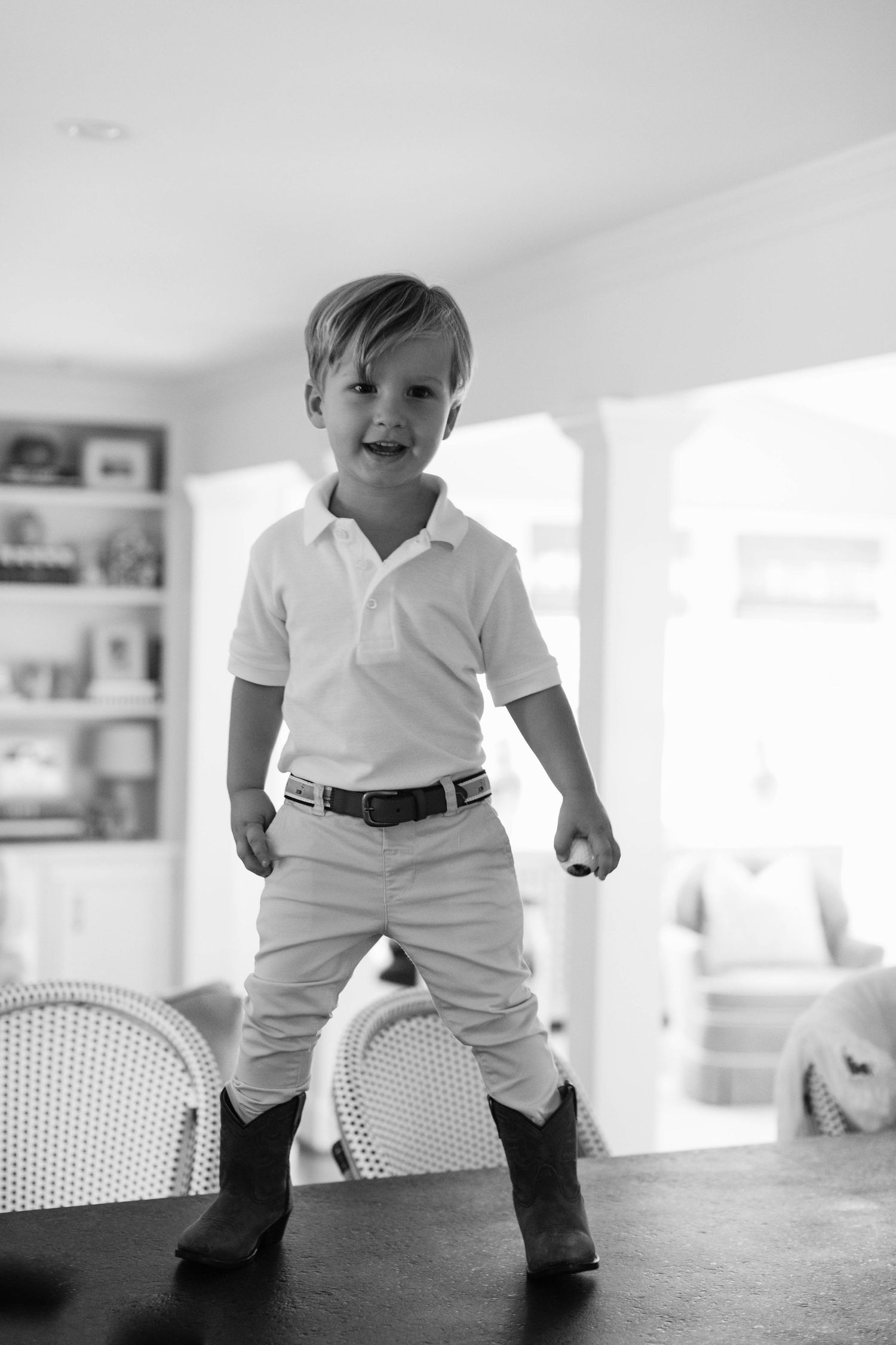 Black and white photo of a smiling child standing on a table wearing a polo shirt, belt, and boots in a bright interior.