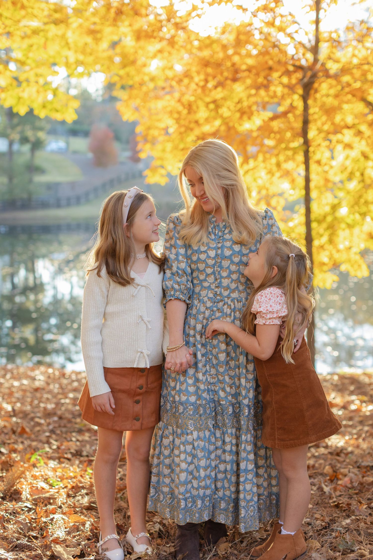 Three young girls in autumn outfits stand together among fallen yellow leaves during a fall photo session.
