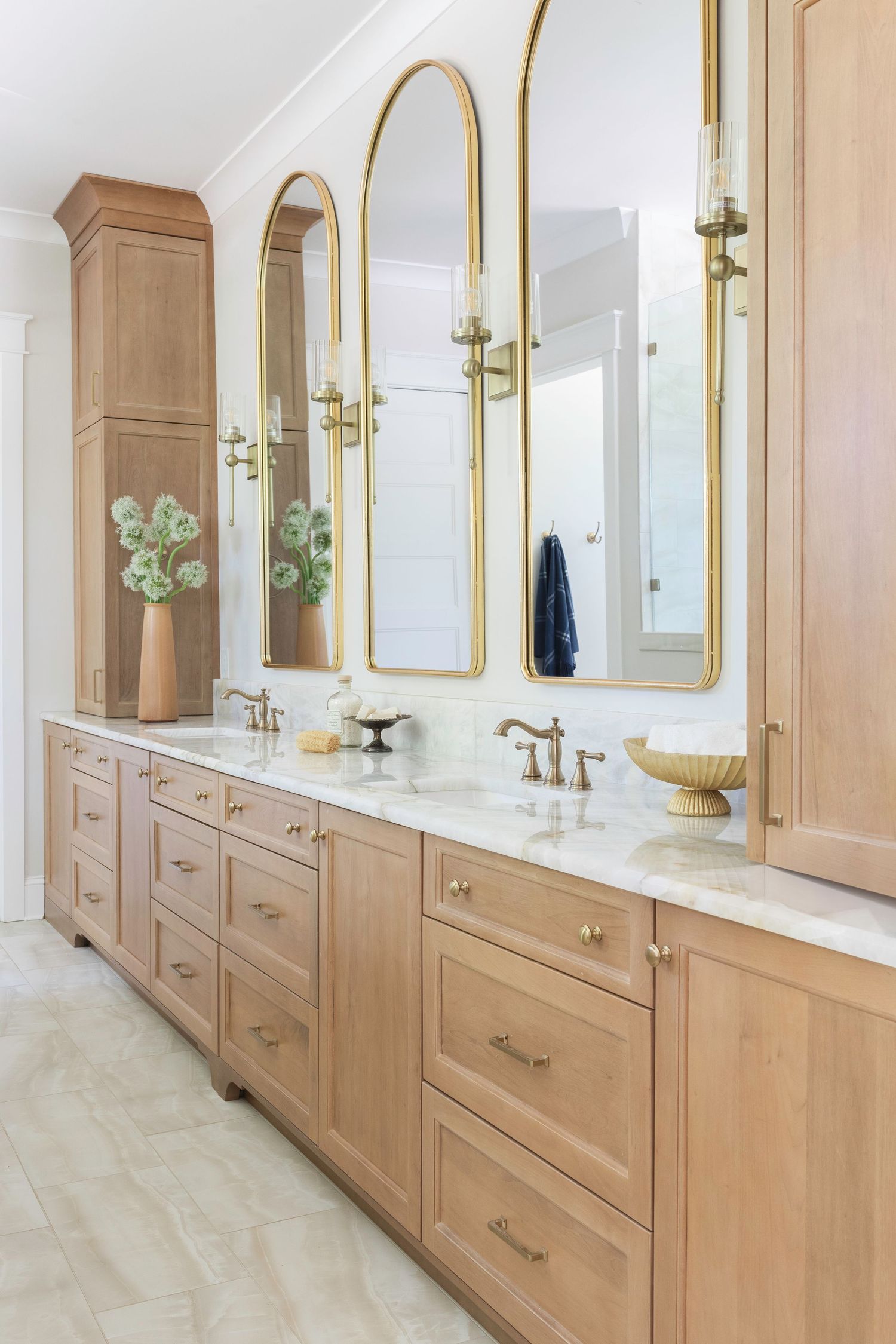 Elegant double vanity bathroom with warm wood cabinets, gold arched mirrors, marble countertops, and brass fixtures.