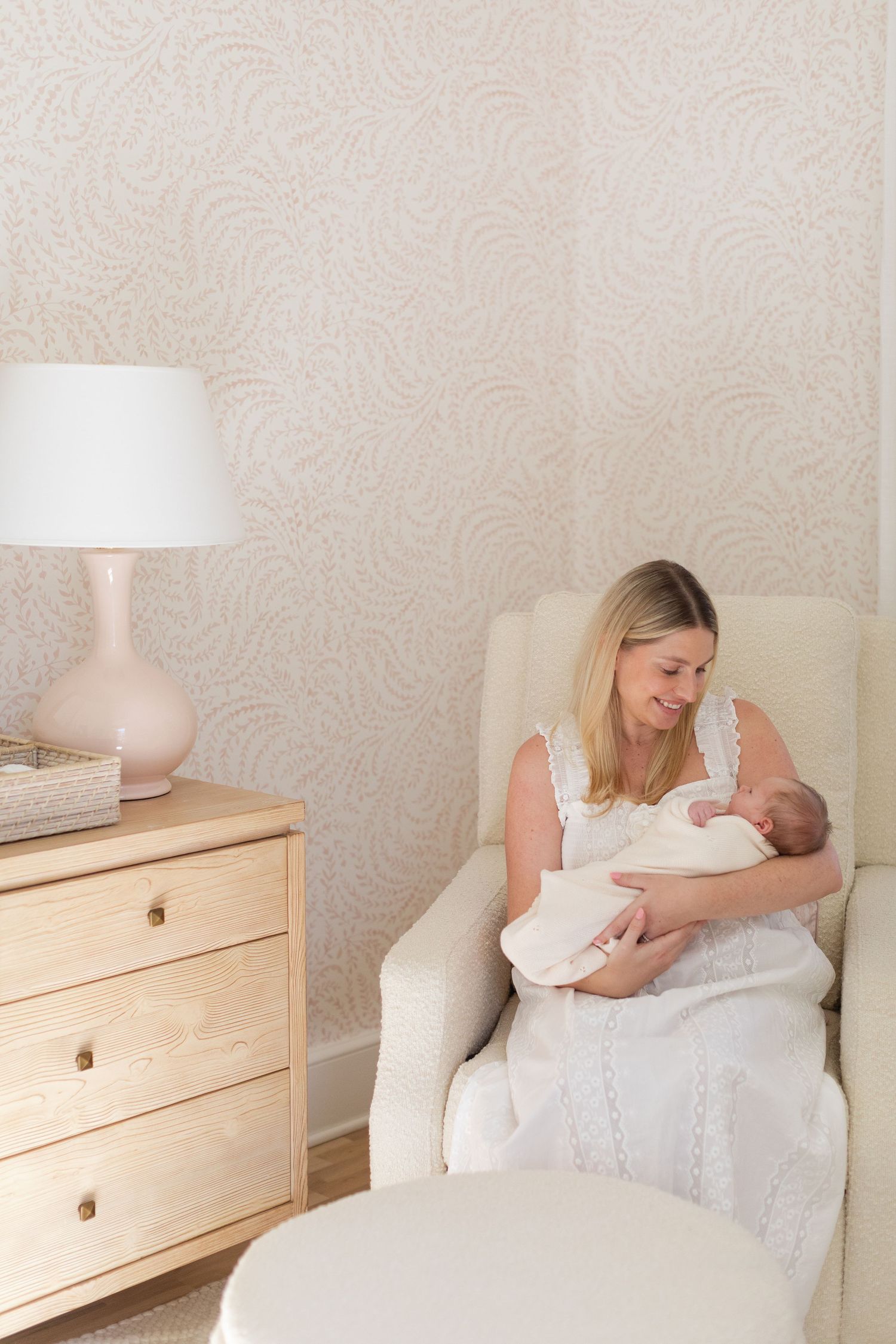 A tender moment in a nursery with a cream armchair, pink table lamp, and wooden dresser creating a peaceful atmosphere.