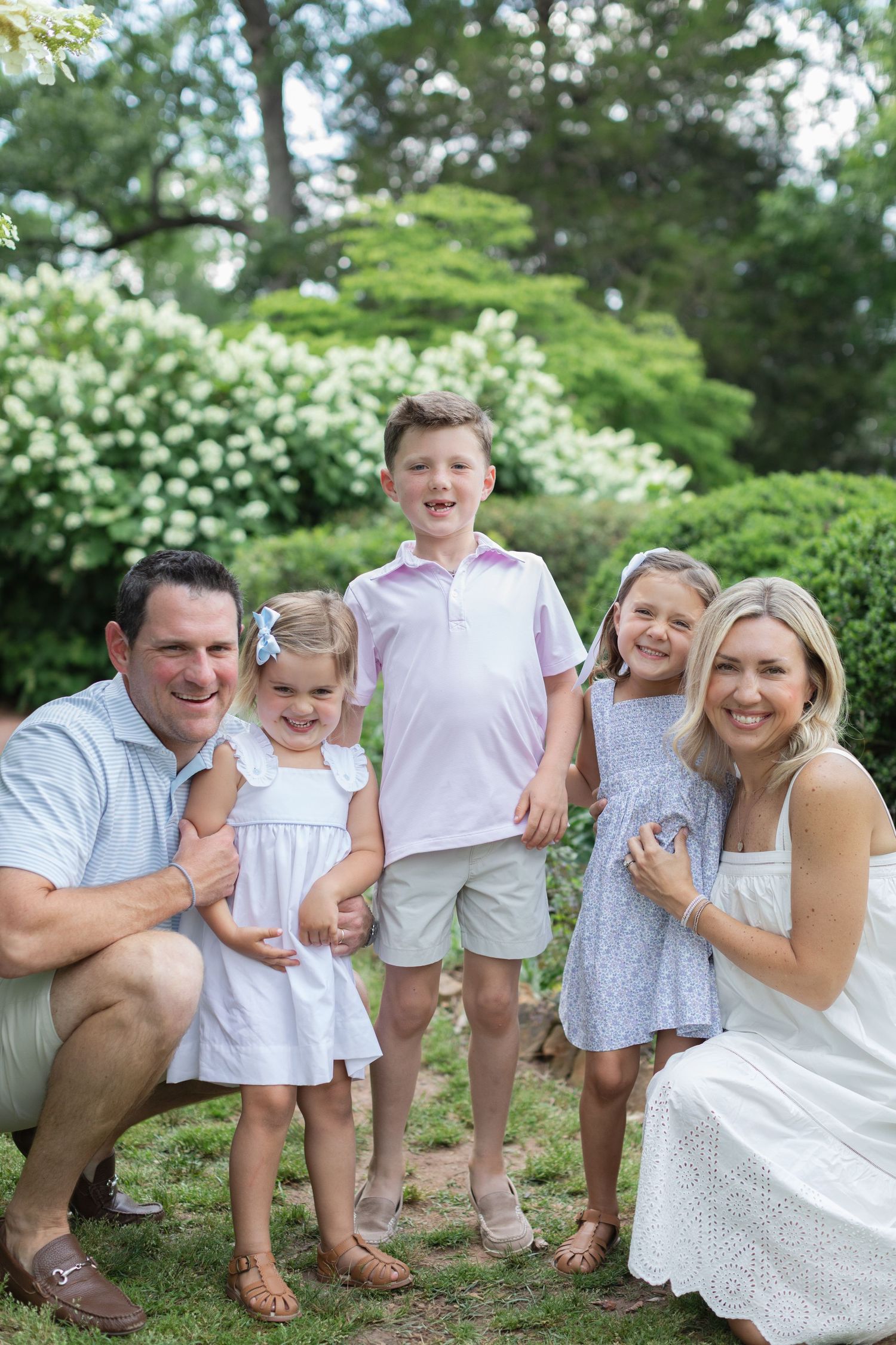 A happy family of five poses outdoors in a lush green garden, wearing coordinated pastel outfits for a portrait.