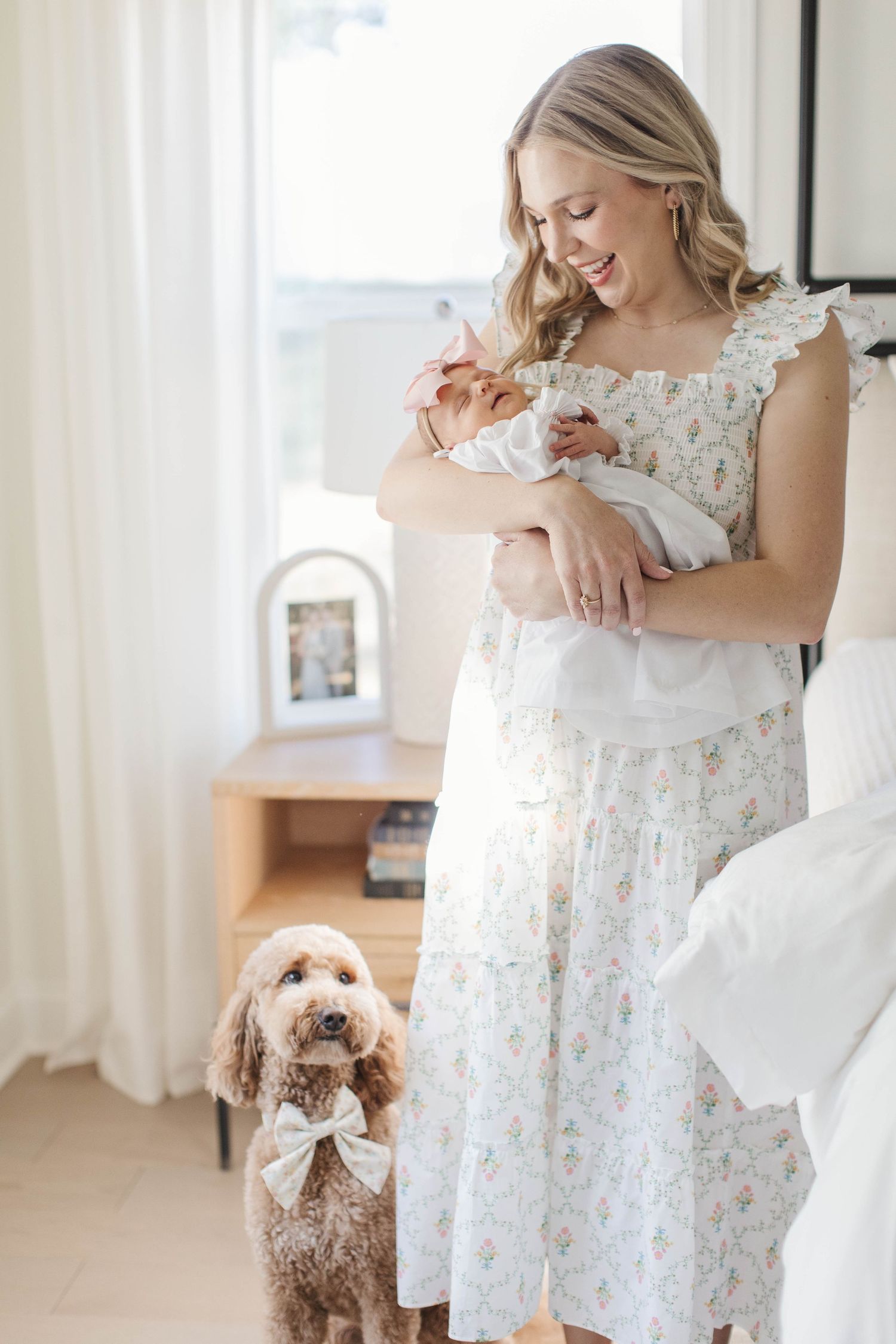 A mother in a floral dress holds a newborn baby while a brown goldendoodle dog with a bow tie stands nearby.