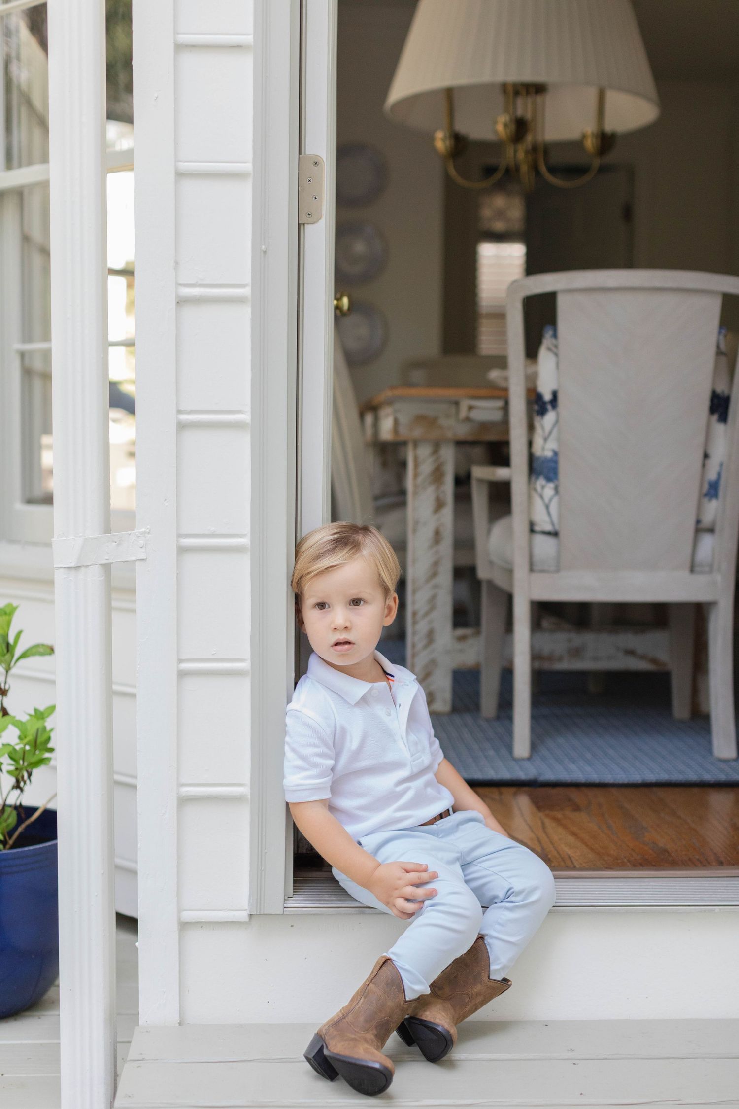 Child in blue outfit sits and plays on a white covered porch with a view into the home.