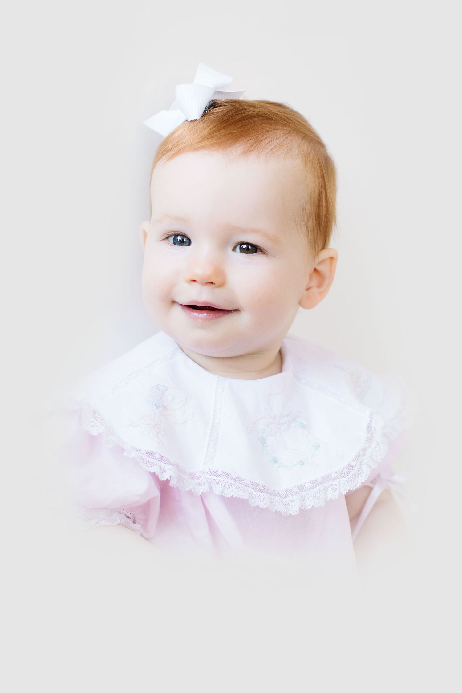 Sweet baby wearing a white lace bib and pink dress with a white bow smiles brightly against light background.