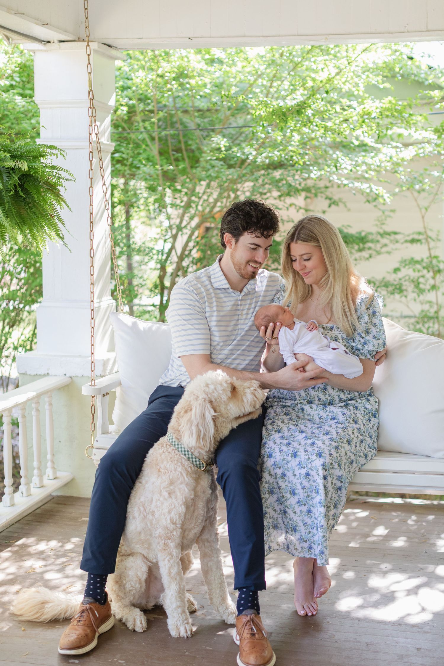 New parents sitting on porch swing with Goldendoodle dog, mother holding newborn baby in white outfit outdoors.