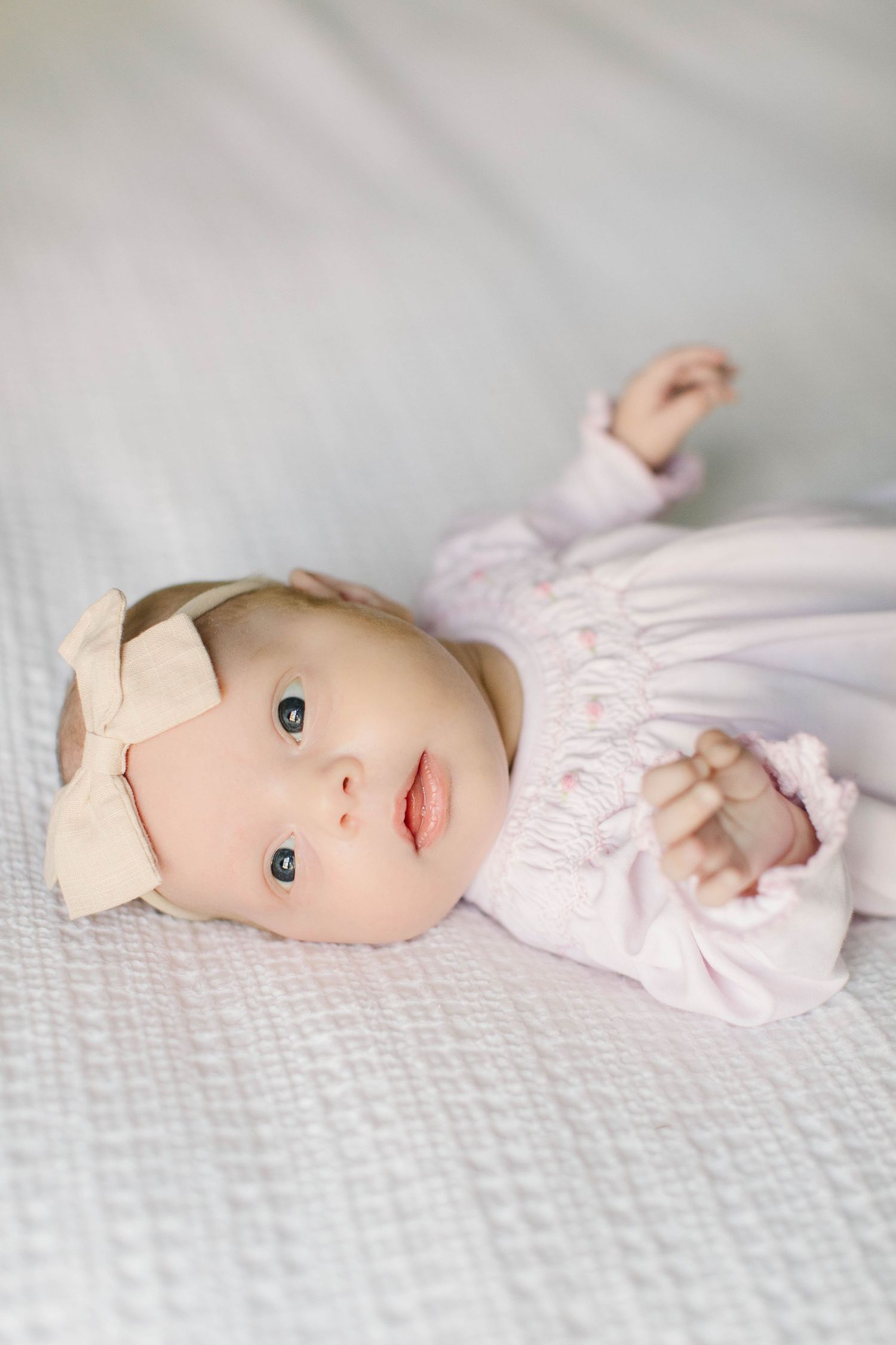 Baby in pink dress lying on textured white bedding with bow hair accessory.