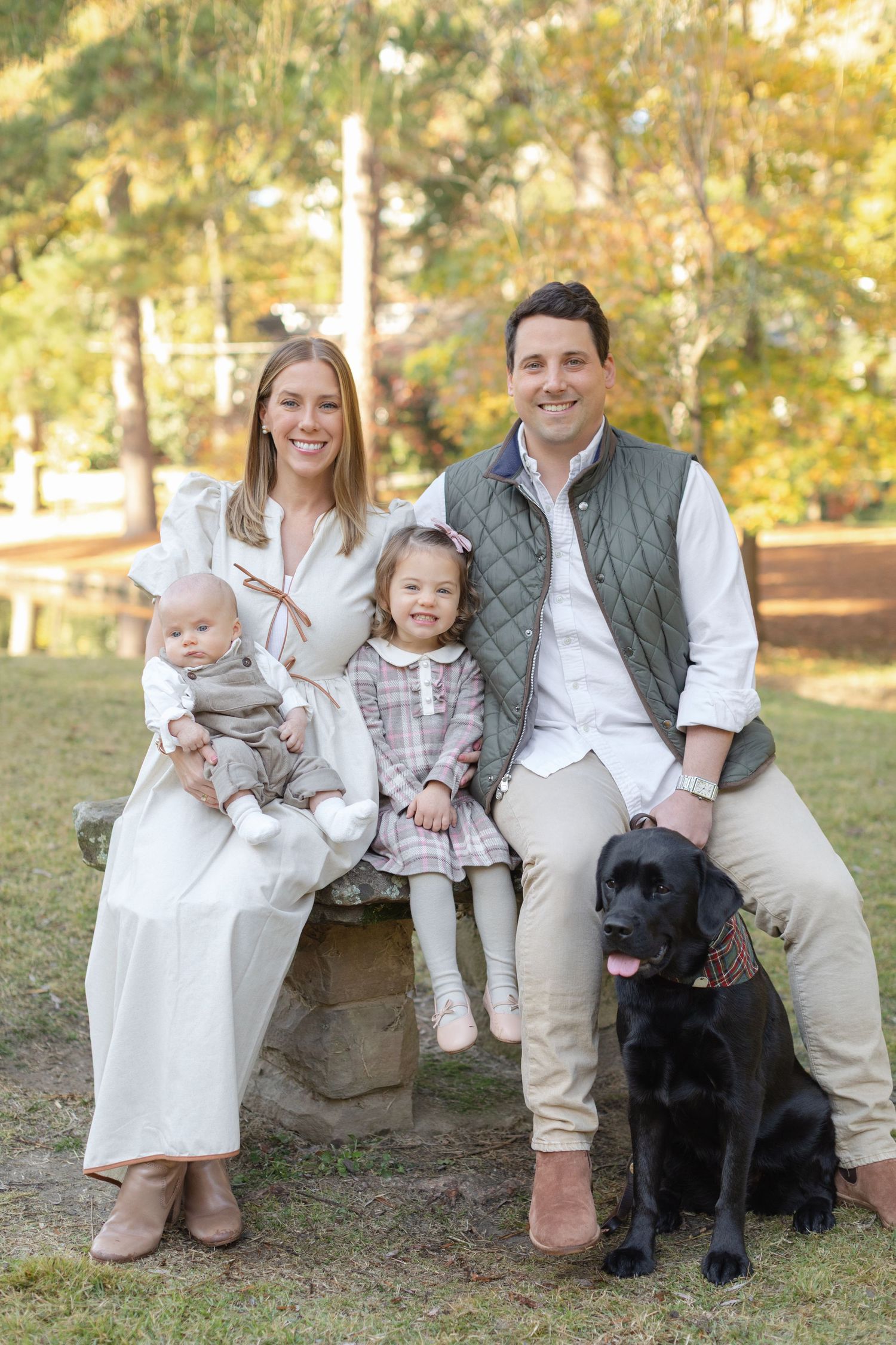 A family poses outdoors in coordinated white and beige outfits with their black Labrador dog on a fall afternoon.